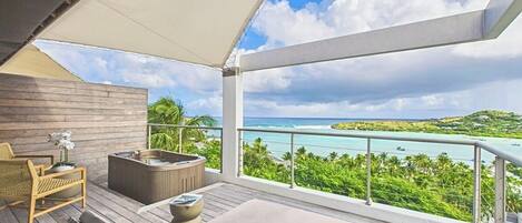 A balcony with a wooden floor, a sofa, a chair, a hot tub, and a view of a turquoise sea and green landscape.