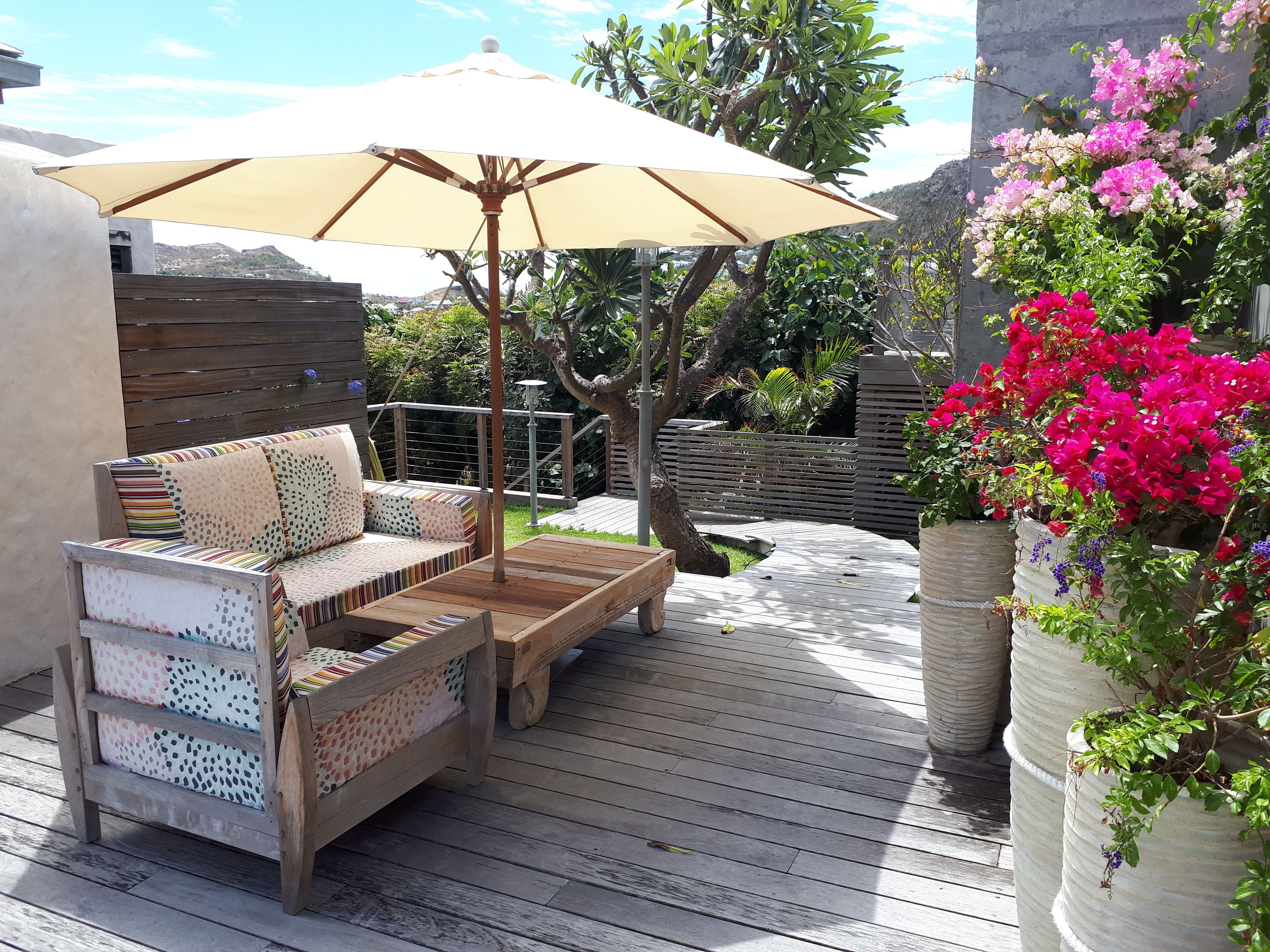 A wooden deck with a sofa and a table, a large umbrella, and potted plants with flowers.