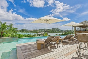 A wooden deck with lounge chairs and umbrellas overlooking a pool and a scenic view of a coastal area. - Amnios St Barth (St. Barthelemy)