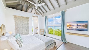 A bedroom with a large bed, a ceiling fan, a view of the sea, and a painting on the wall. - Amnios St Barth (St. Barthelemy)