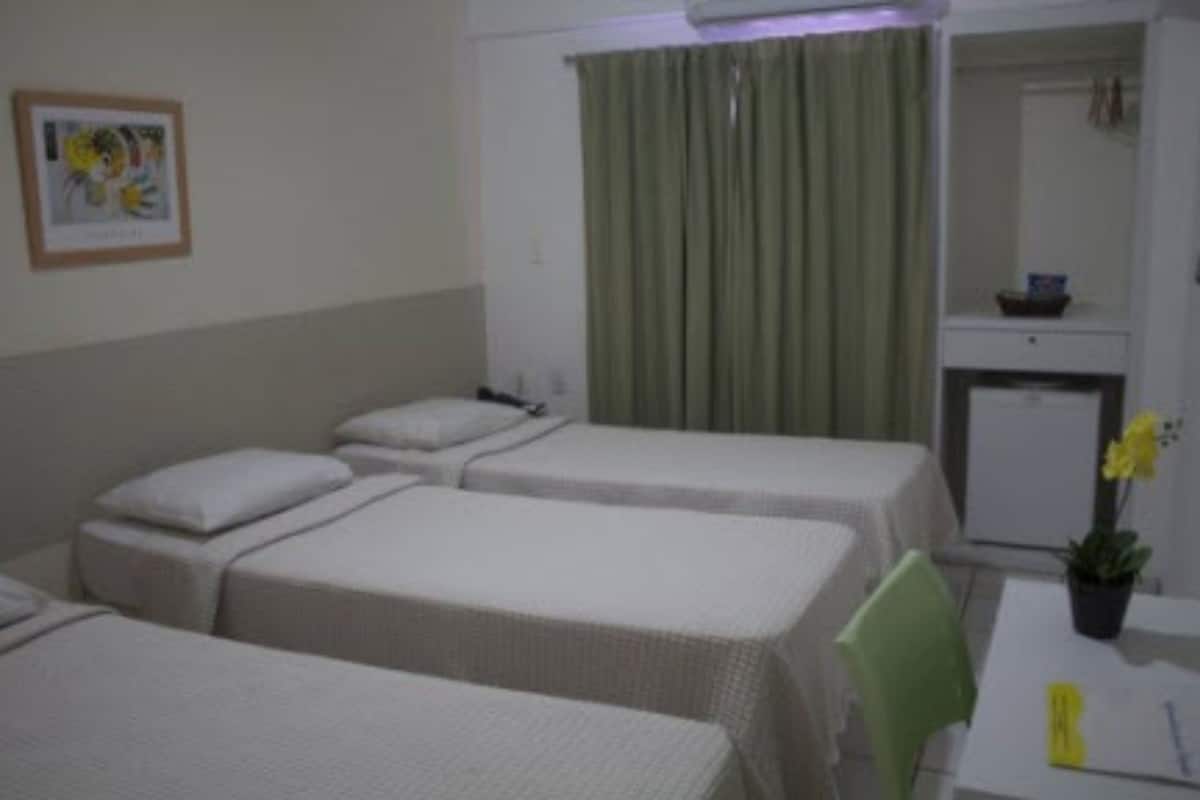 Quarto de hotel com duas camas de solteiro, uma mesinha com cadeira, escrivaninha, planta em vaso e espelho sobre a escrivaninha.