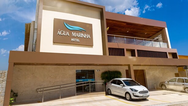 The exterior of the Água Marinha Hotel with a modern design, featuring a prominent sign, a covered parking area, and a clear blue sky in the background. - Hotel Água Marinha (Natal)
