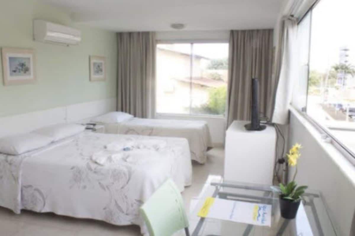 Quarto de hotel com duas camas, uma mesa pequena com uma cadeira, um vaso de planta e uma janela com cortinas.