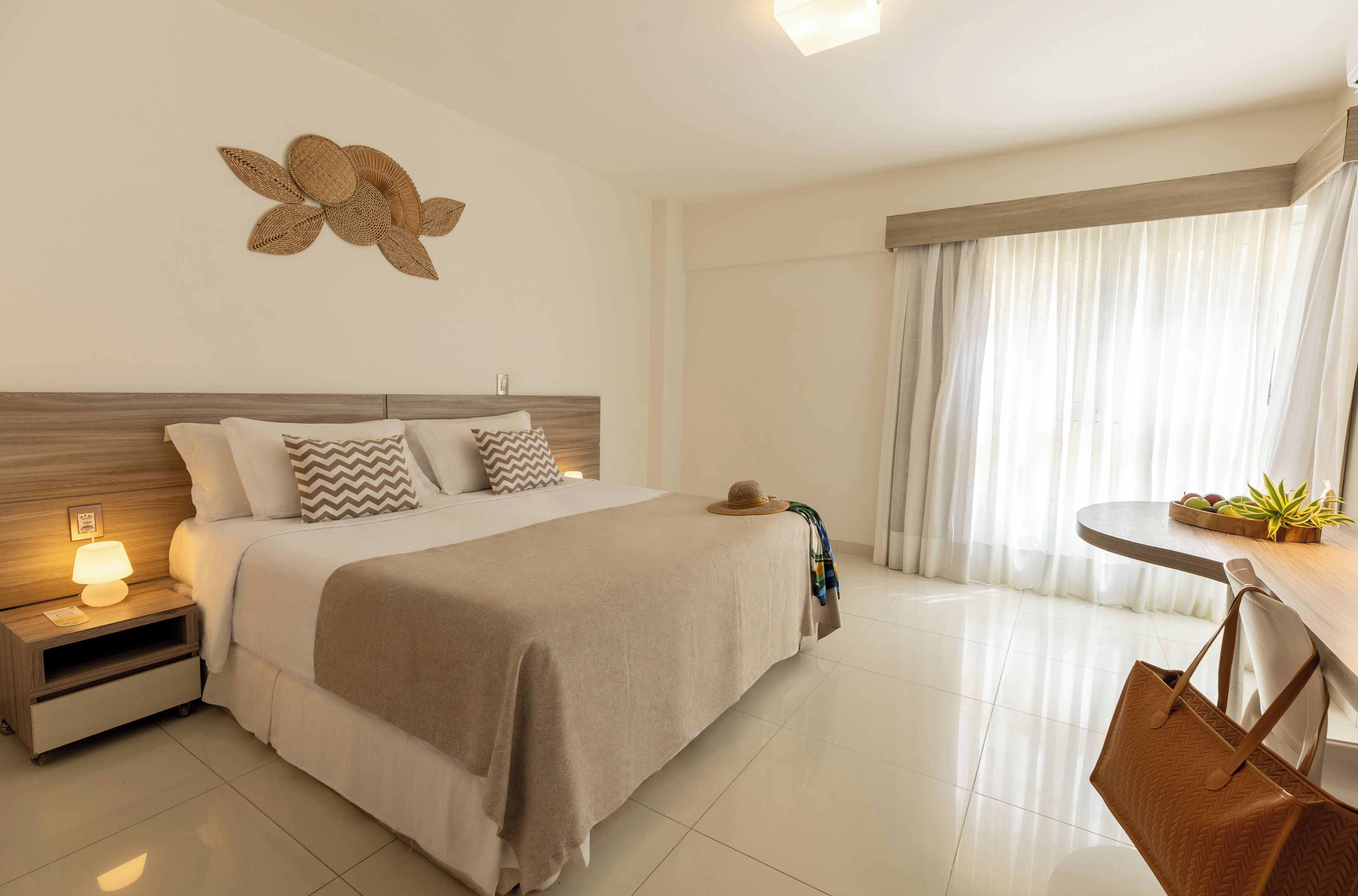Habitación de hotel con cama, mesita de noche, lámpara y vistas al exterior a través de cortinas.