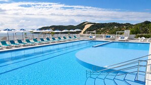 A large swimming pool with multiple lounge chairs and umbrellas, surrounded by a white fence and a view of a hill. - SERHS Beach Hotel (Natal)