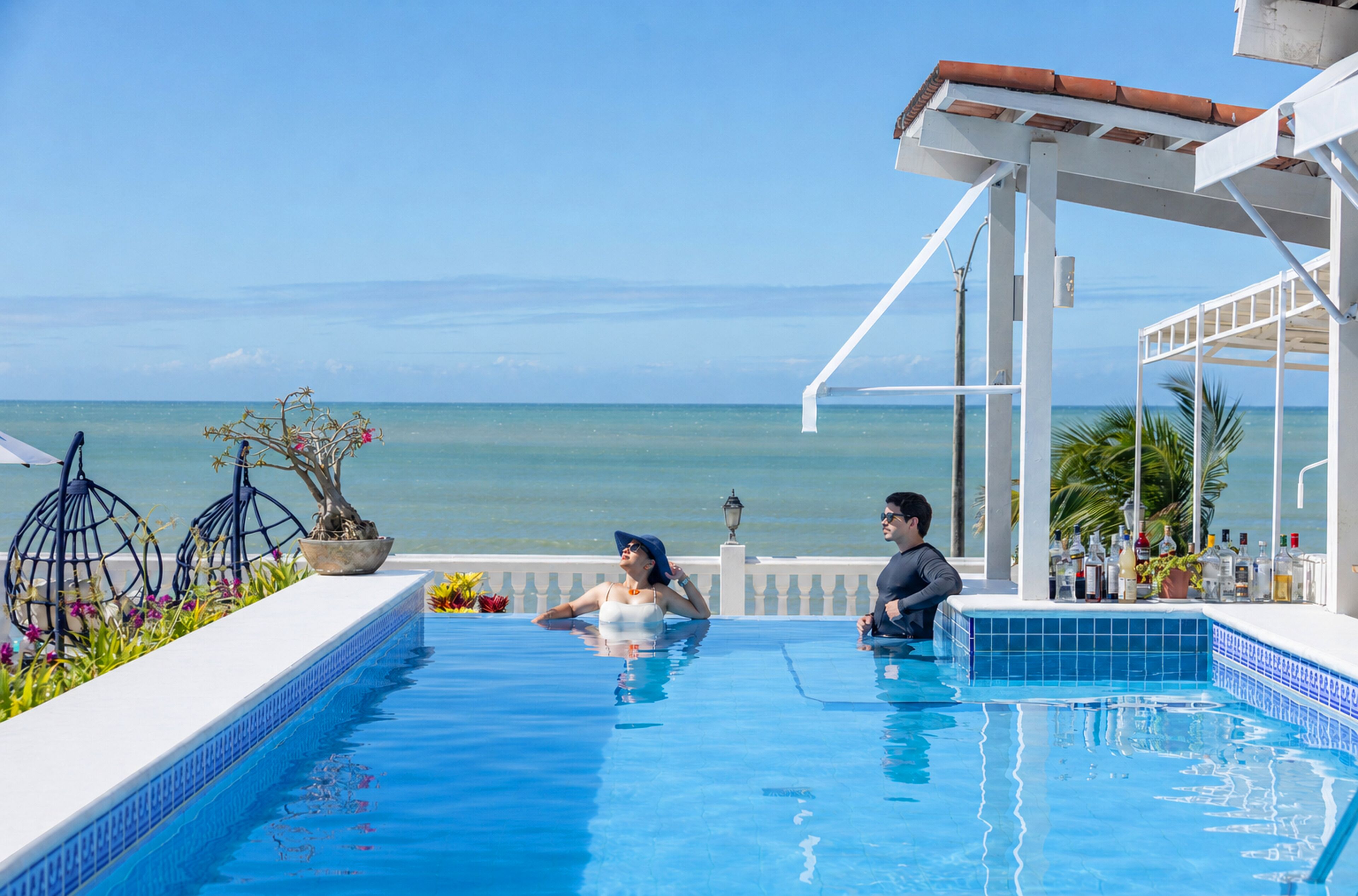A poolside area with a view of the ocean, a covered seating area, and a bar with bottles.