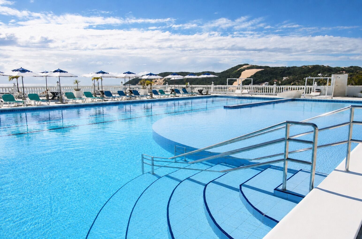 Piscina azul cristalina con sillas de descanso y sombrillas, rodeada por una valla blanca y vistas a las colinas.