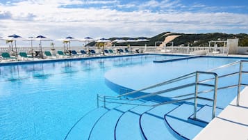 Piscina azul cristalina con sillas de descanso y sombrillas, rodeada por una valla blanca y vistas a las colinas.