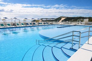 Piscina azul límpida com espreguiçadeiras e guarda-sóis, cercada por um muro branco e vista para colinas.