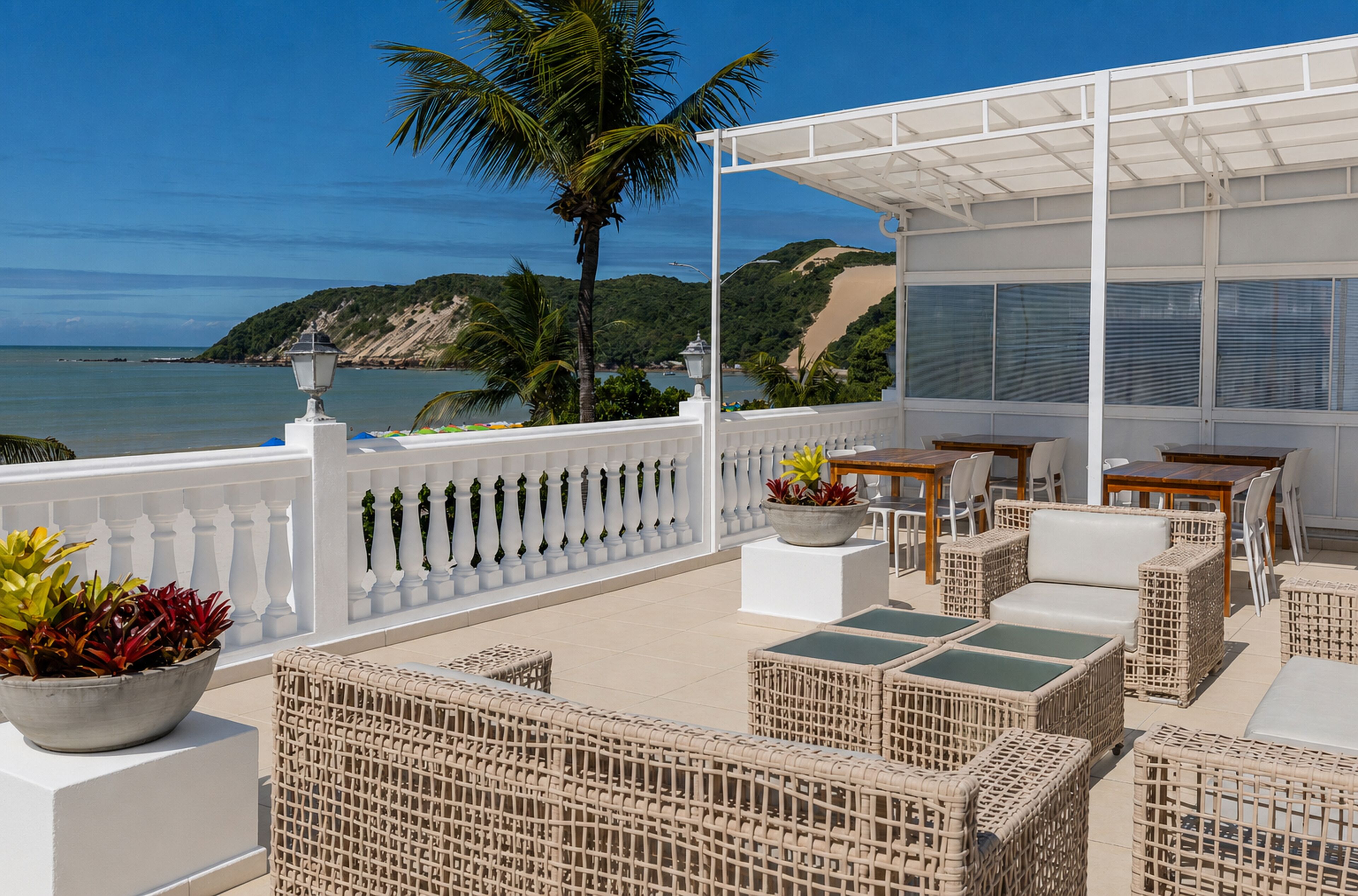Terraço com móveis de vime, guarda-corpo branco e vista para o mar e colinas.