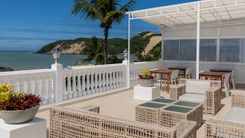 Terraço com móveis de vime, guarda-corpo branco e vista para o mar e colinas.