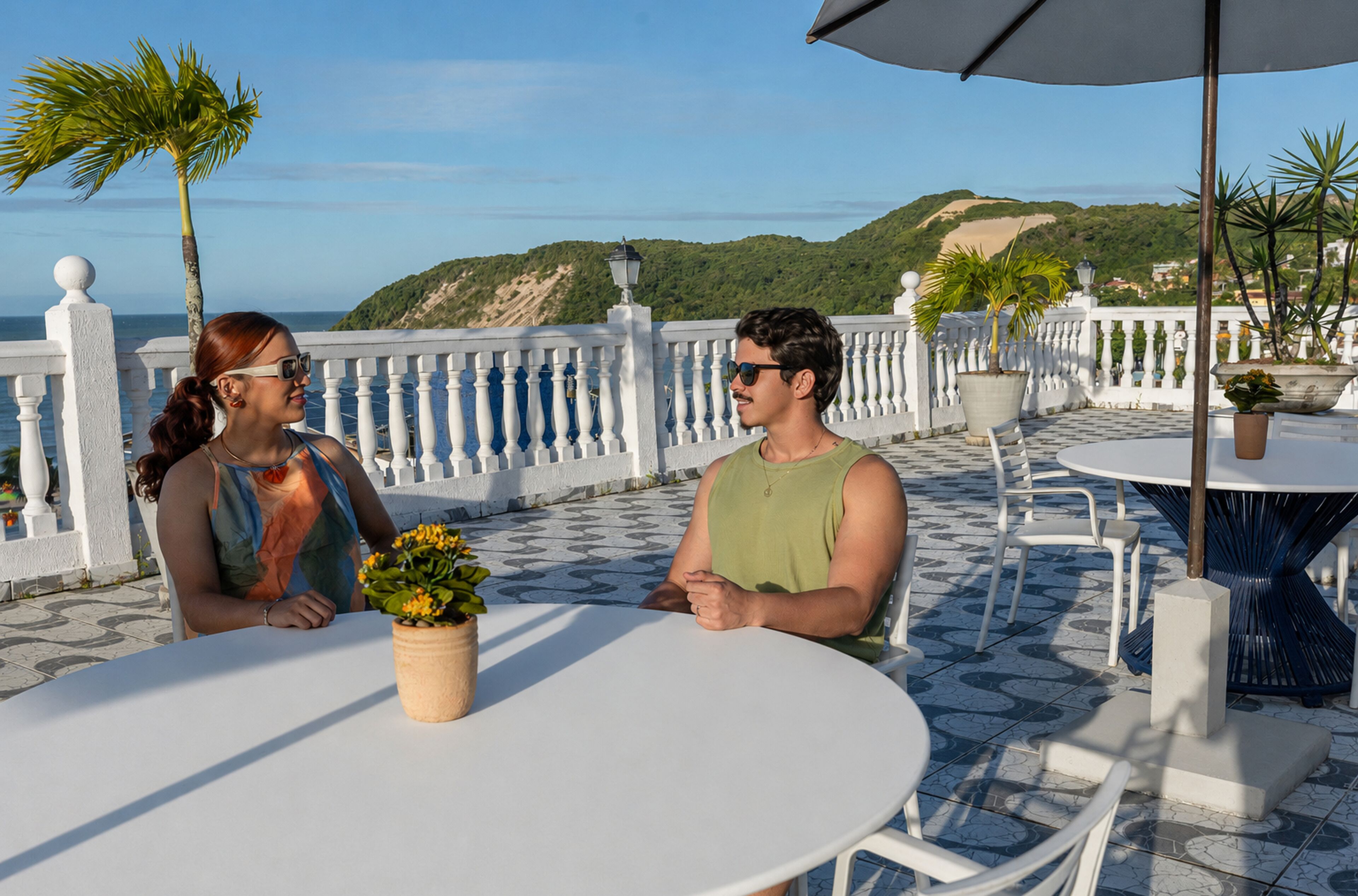 Eine Terrasse mit weißer Brüstung, Sitzmöbeln im Freien und Blick auf Meer und Hügel.