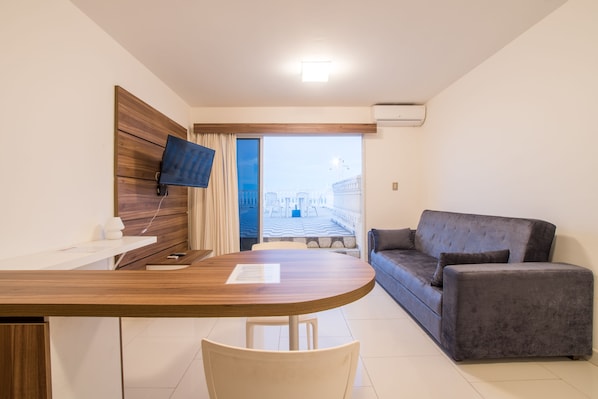Ett modernt hotellrum med ett skrivbord i trä, en platt-TV som är monterad på en träpanel, en grå soffa och utsikt över en stadssiluett genom ett stort fönster.