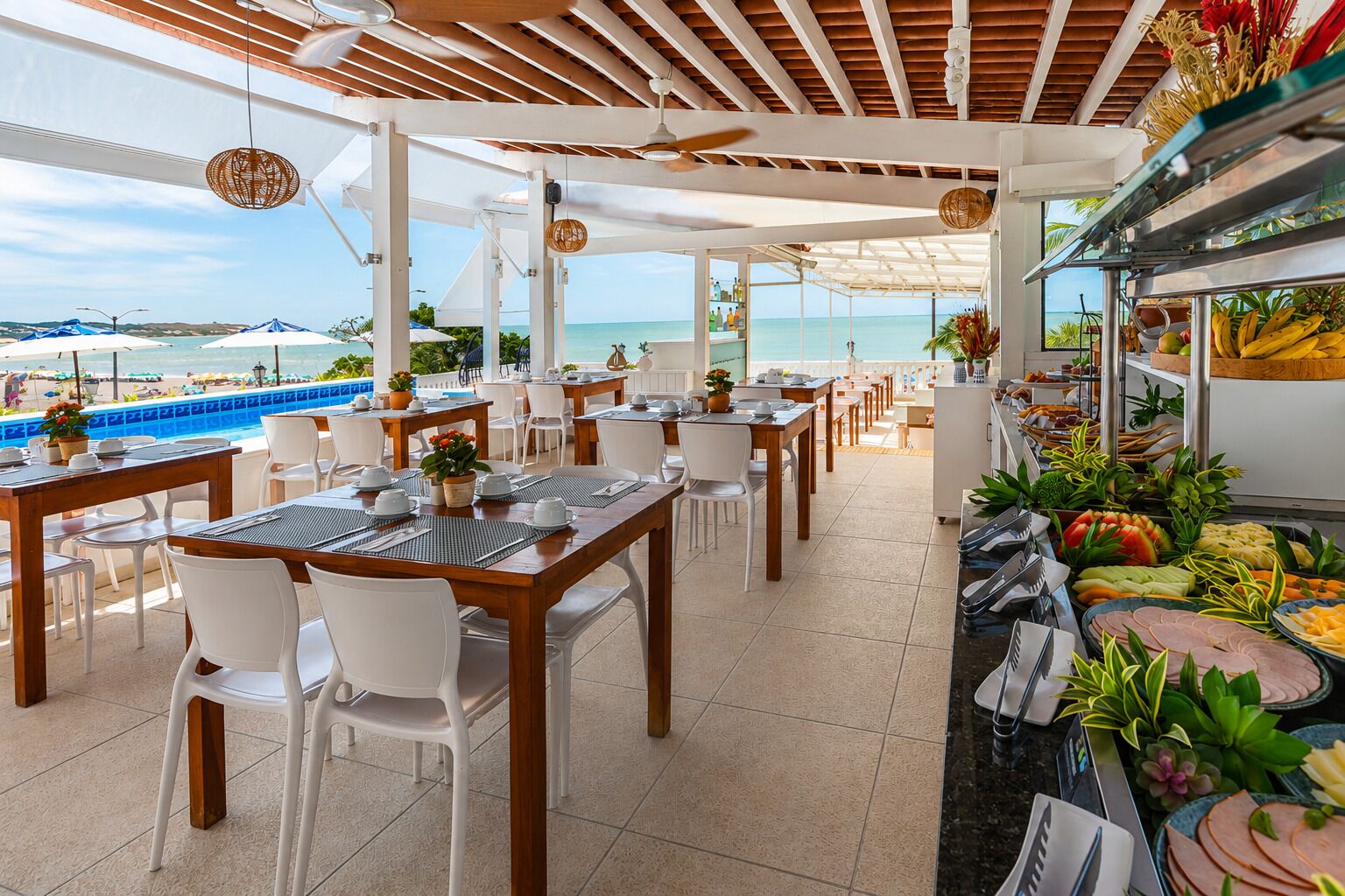 A beachside restaurant with outdoor seating, a buffet with tropical fruits and vegetables, and a view of the ocean.