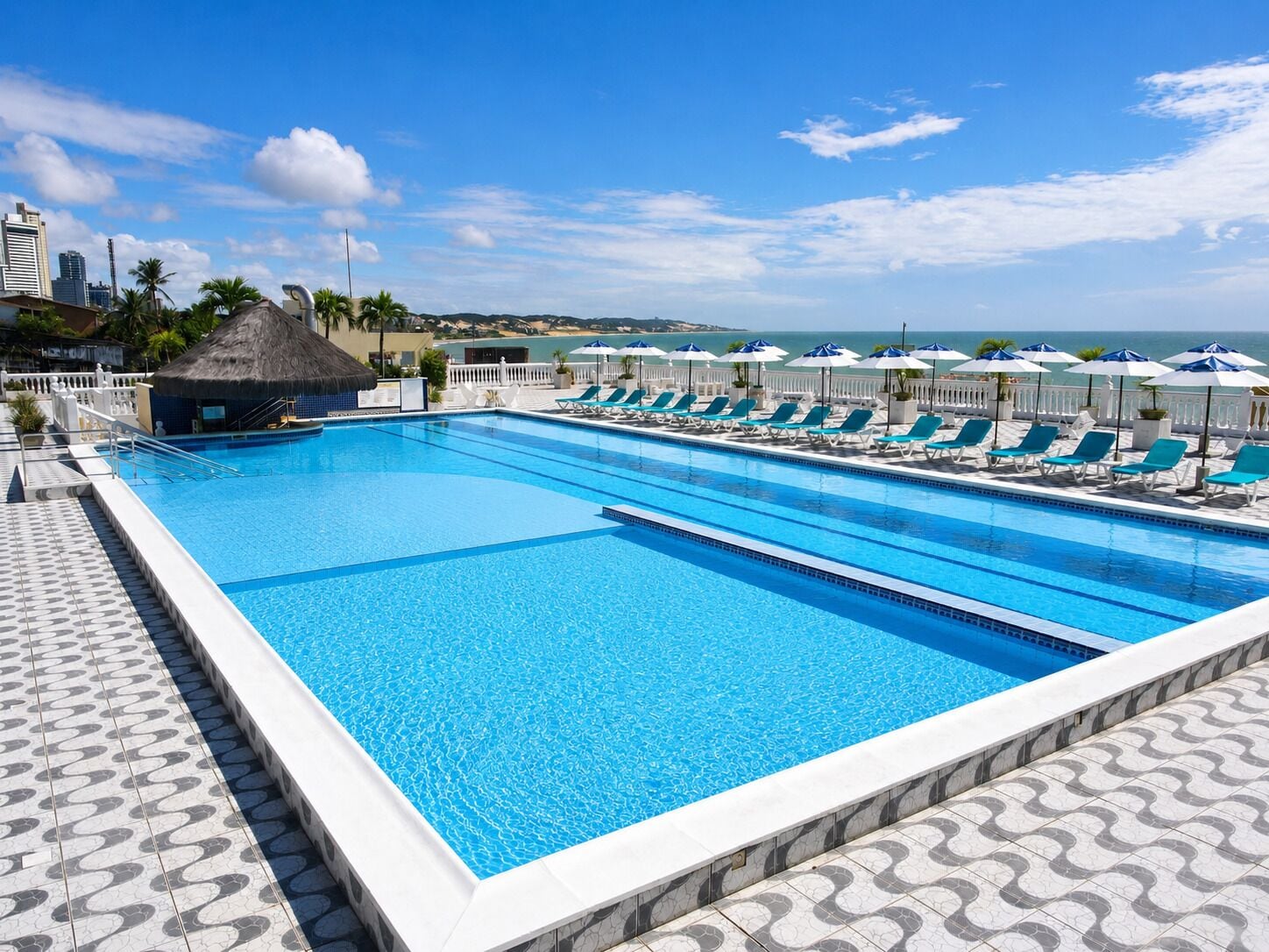 A large rectangular swimming pool with multiple lanes, surrounded by lounge chairs and umbrellas, with a view of the sea and buildings in the background.