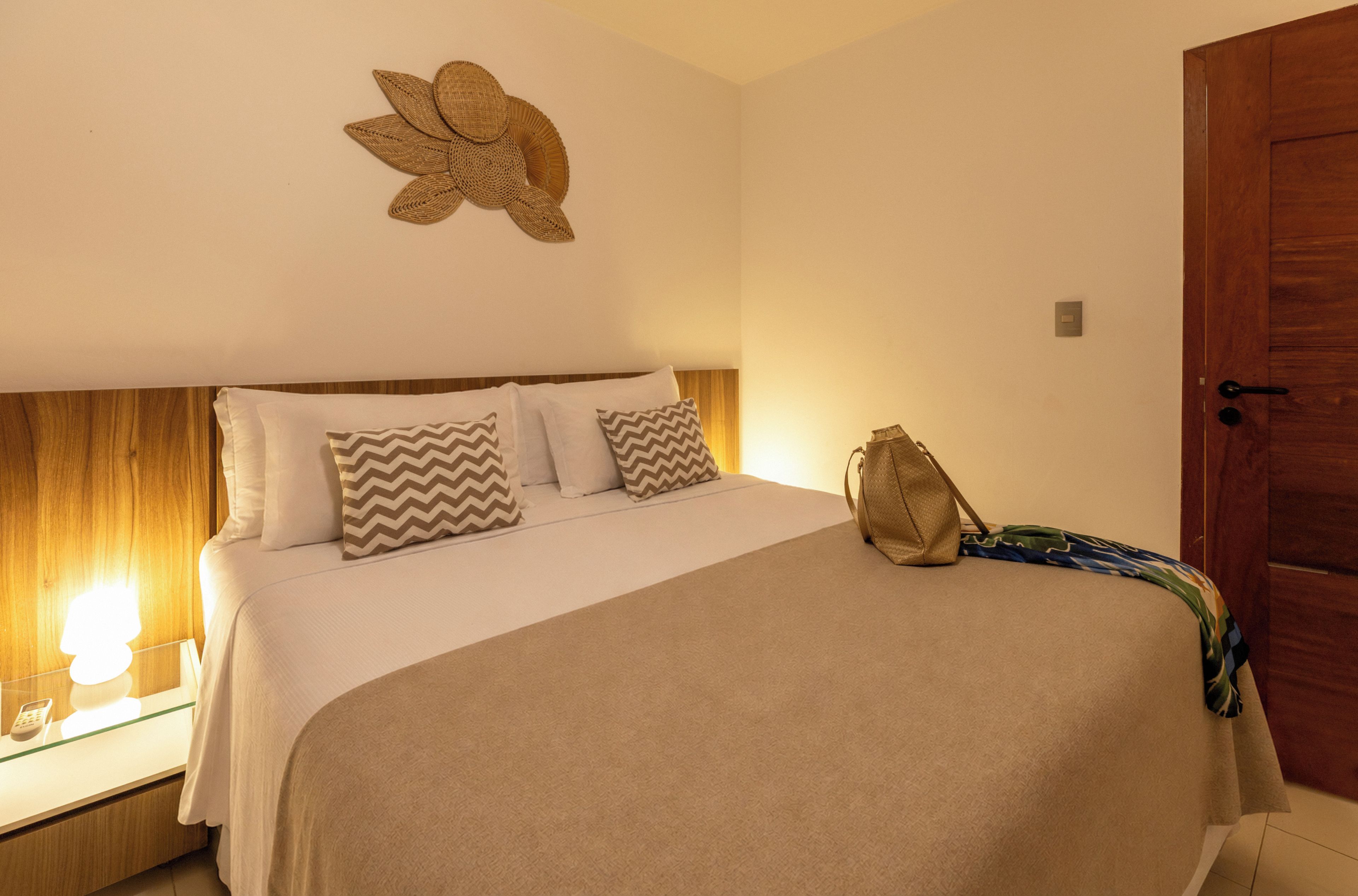 A hotel room with a bed, pillows, a nightstand, a lamp, and a wooden door.