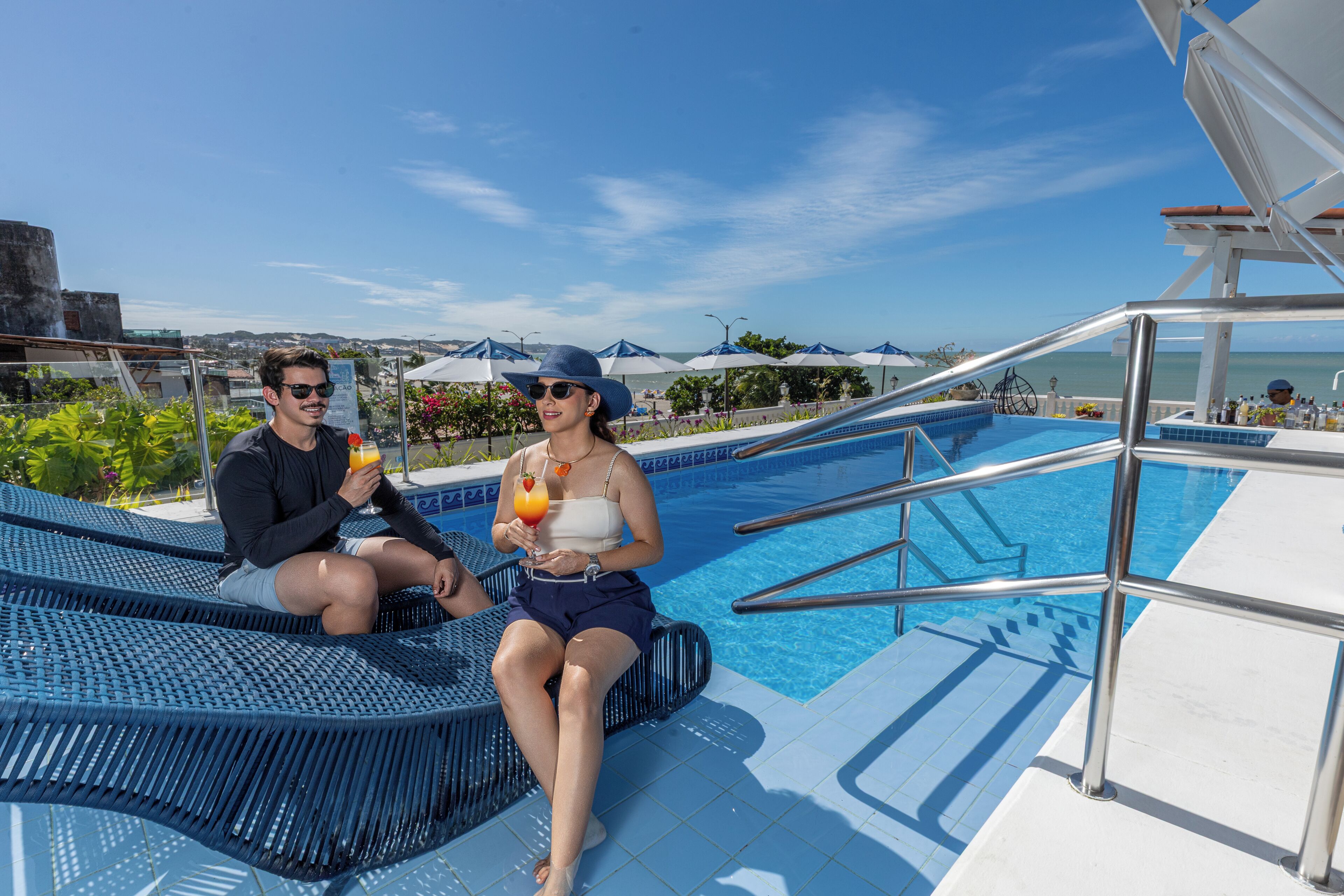 Ein Paar entspannt am Pool mit Blick auf das Meer und eine Stadtlandschaft im Hintergrund.