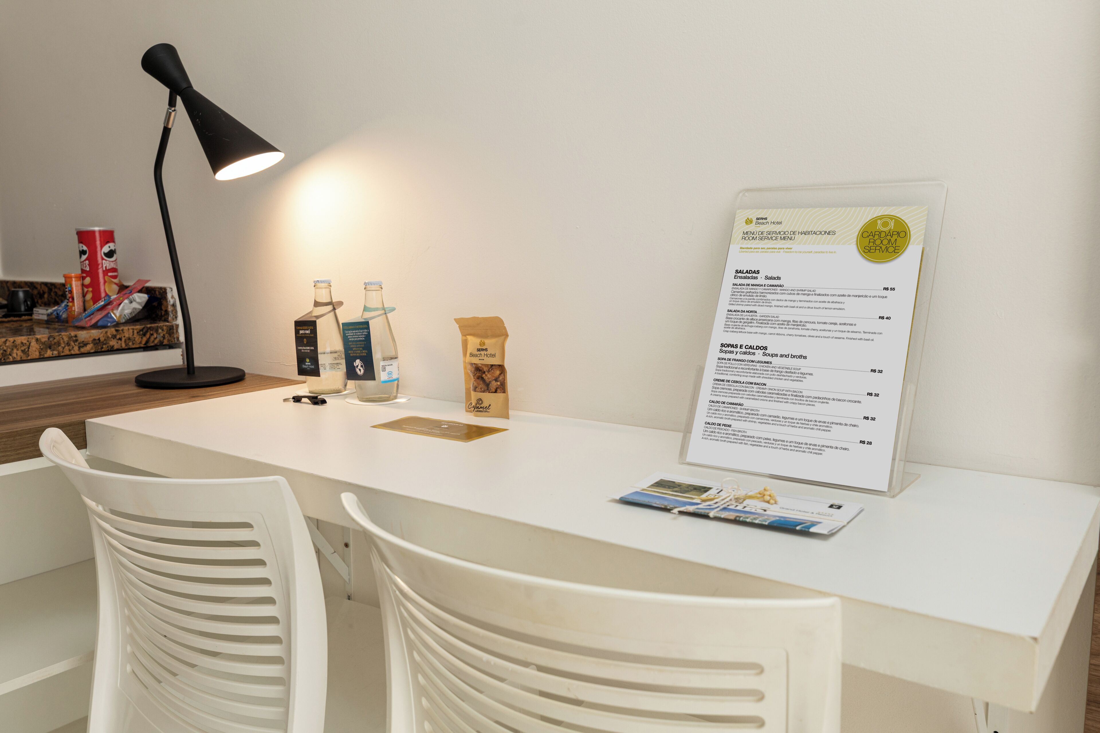 A clean, white hotel room with a desk, two white chairs, a black desk lamp, a menu board, and a small shelf with snacks.