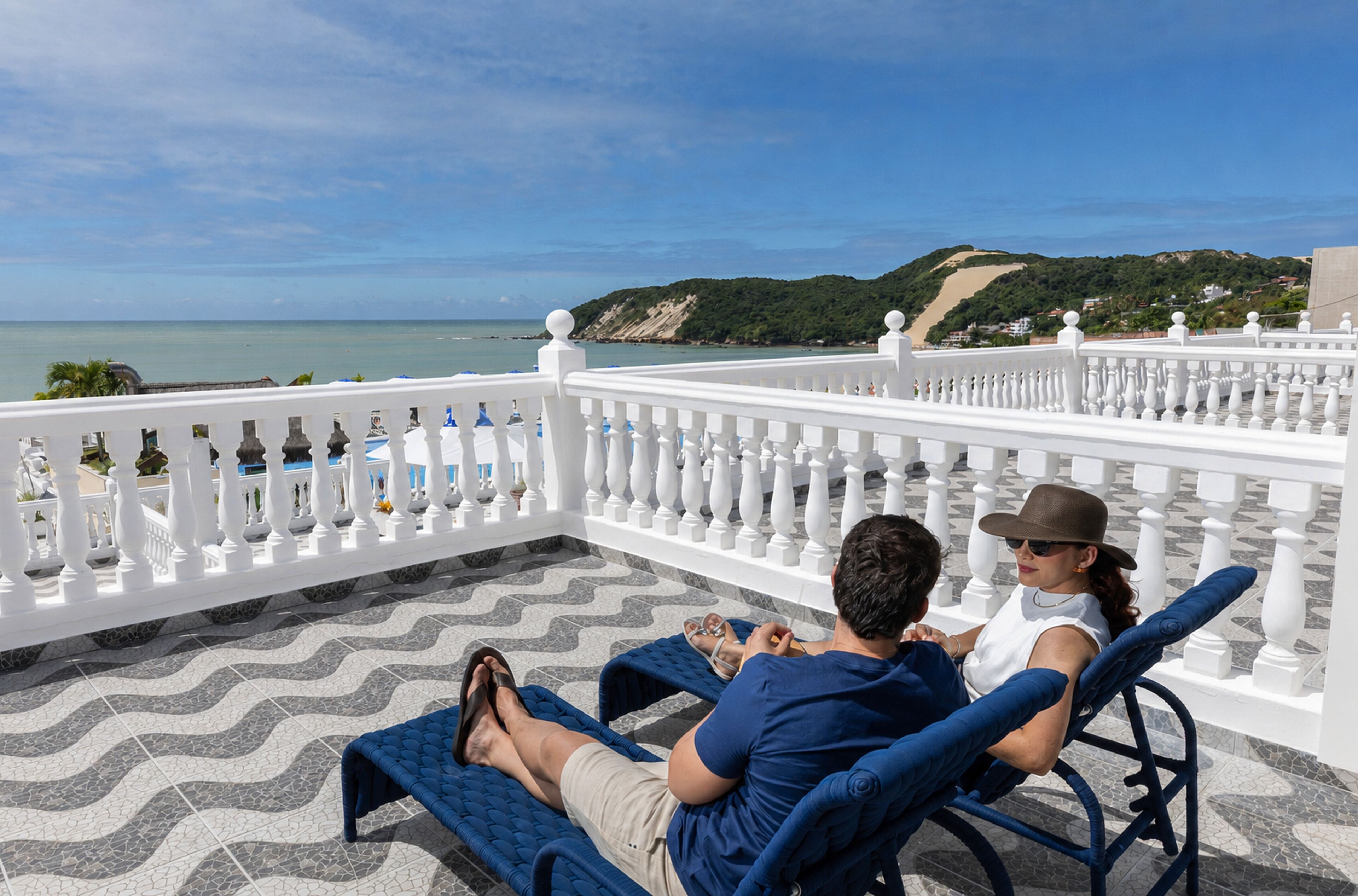 A couple relaxing on a sun lounger with a view of the sea and hills.