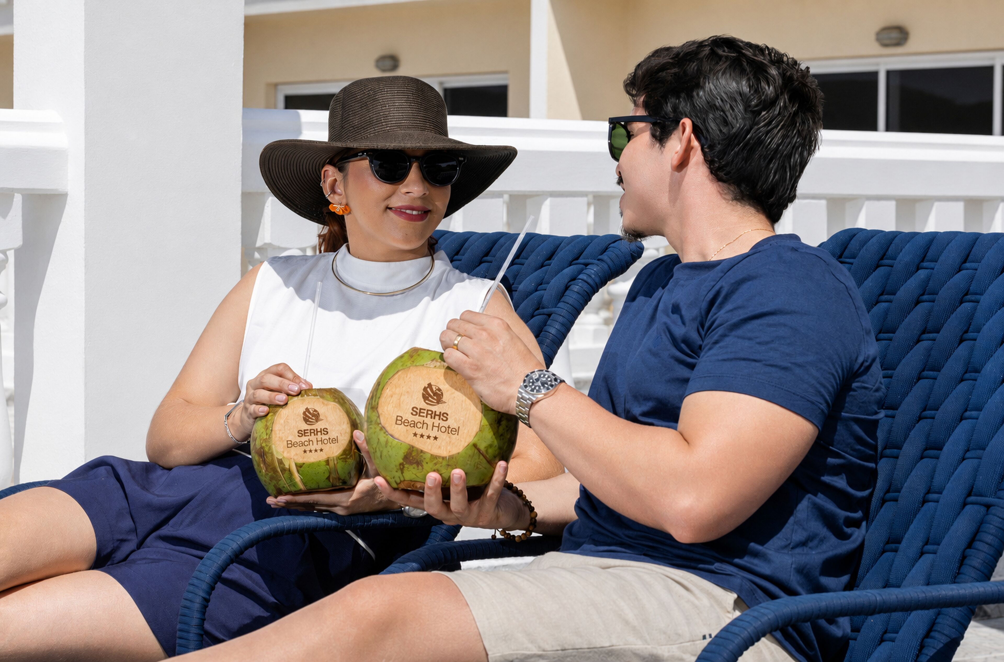 Dos personas sentadas en una silla de lounge azul al aire libre, sosteniendo dos cocos con el logo del SERHS Beach Hotel.