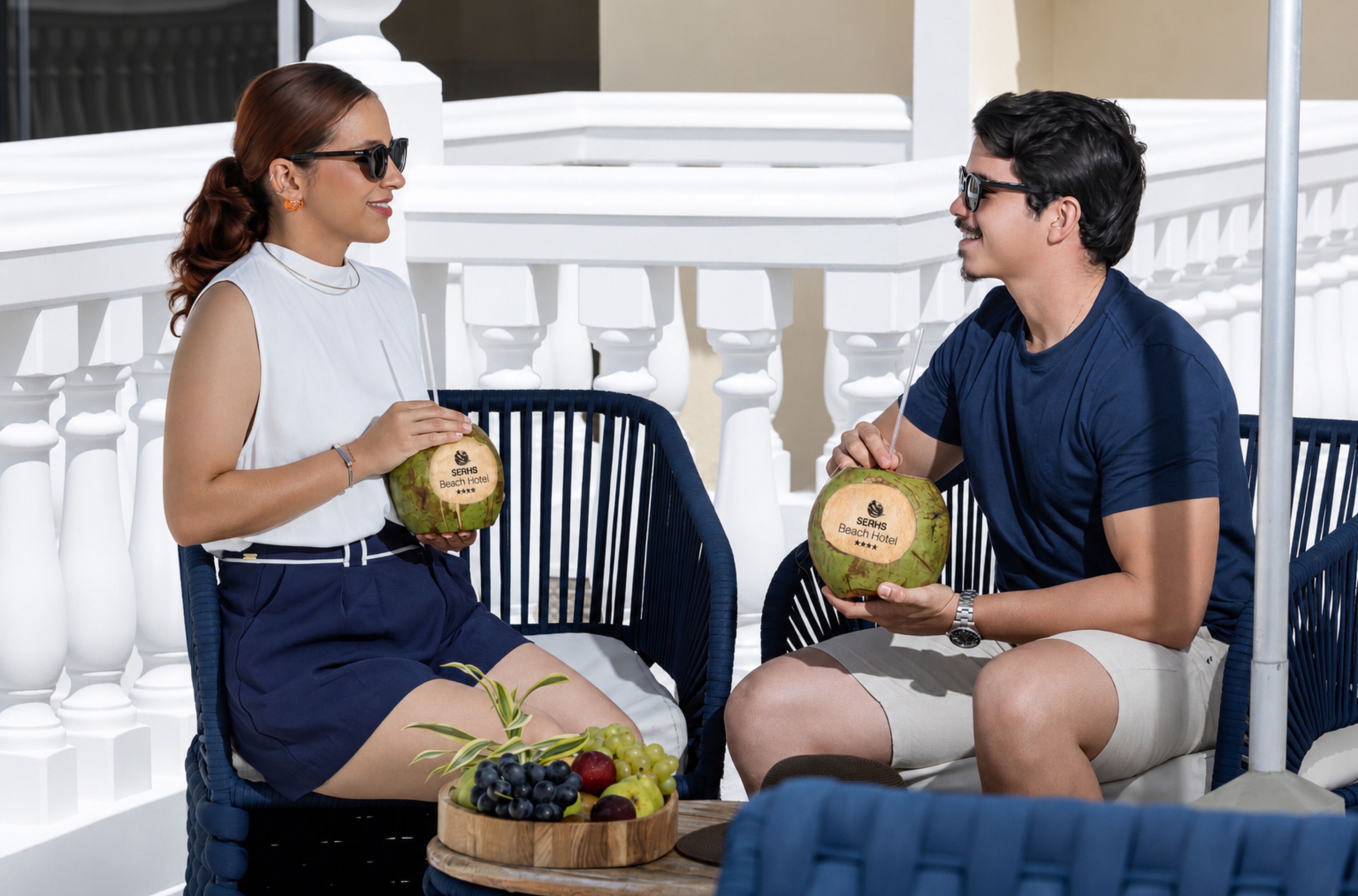 Two people seated on a balcony with a white railing, holding coconuts with a hotel logo, and a fruit bowl with grapes and other fruits in front of them.