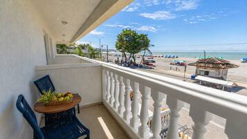 A balcony with a table and chairs, overlooking a beach with umbrellas and a clear sky.