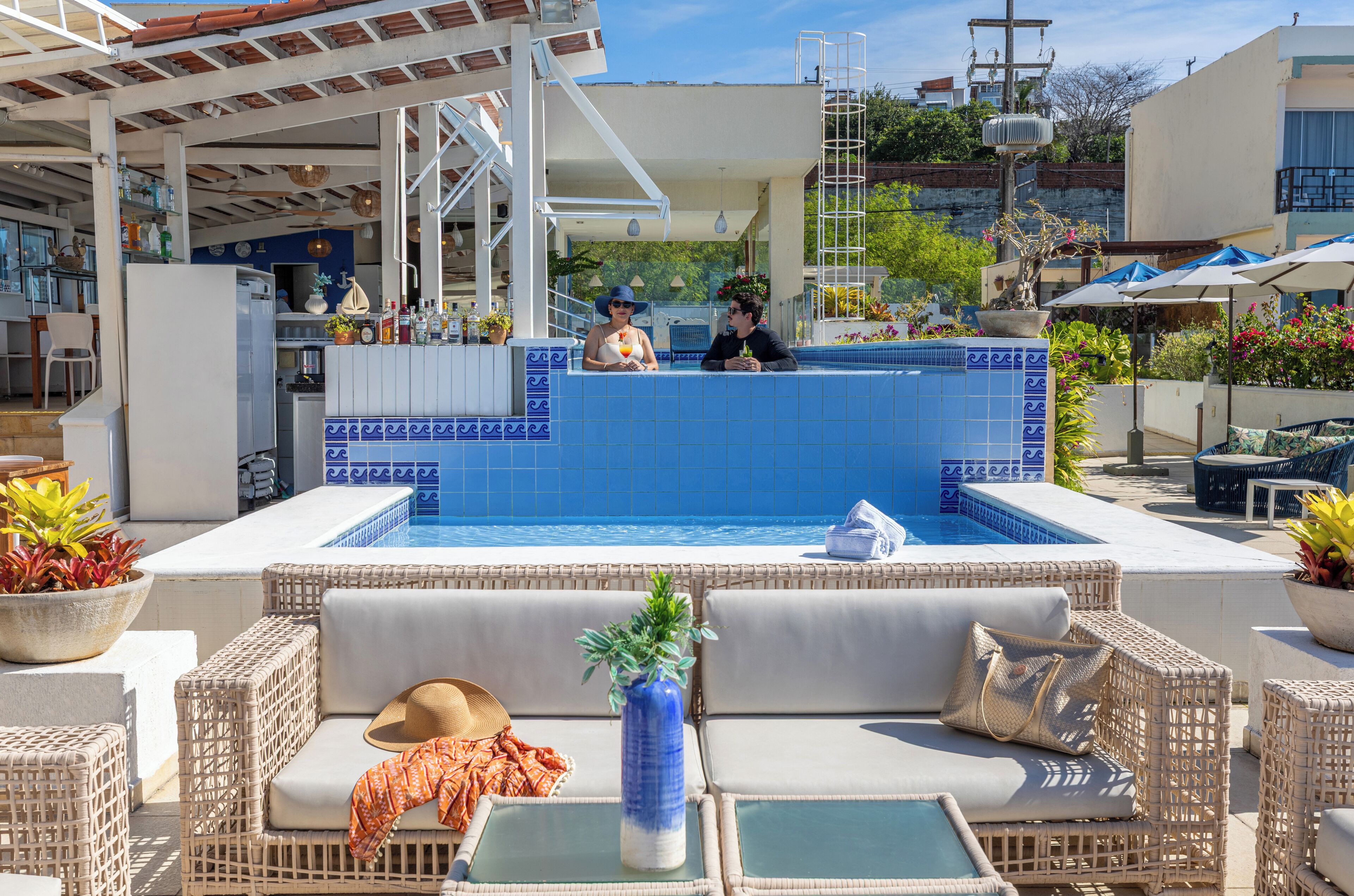 Zona de piscina con pared de azulejos azules, mobiliario de mimbre y una barra con botellas y una planta.