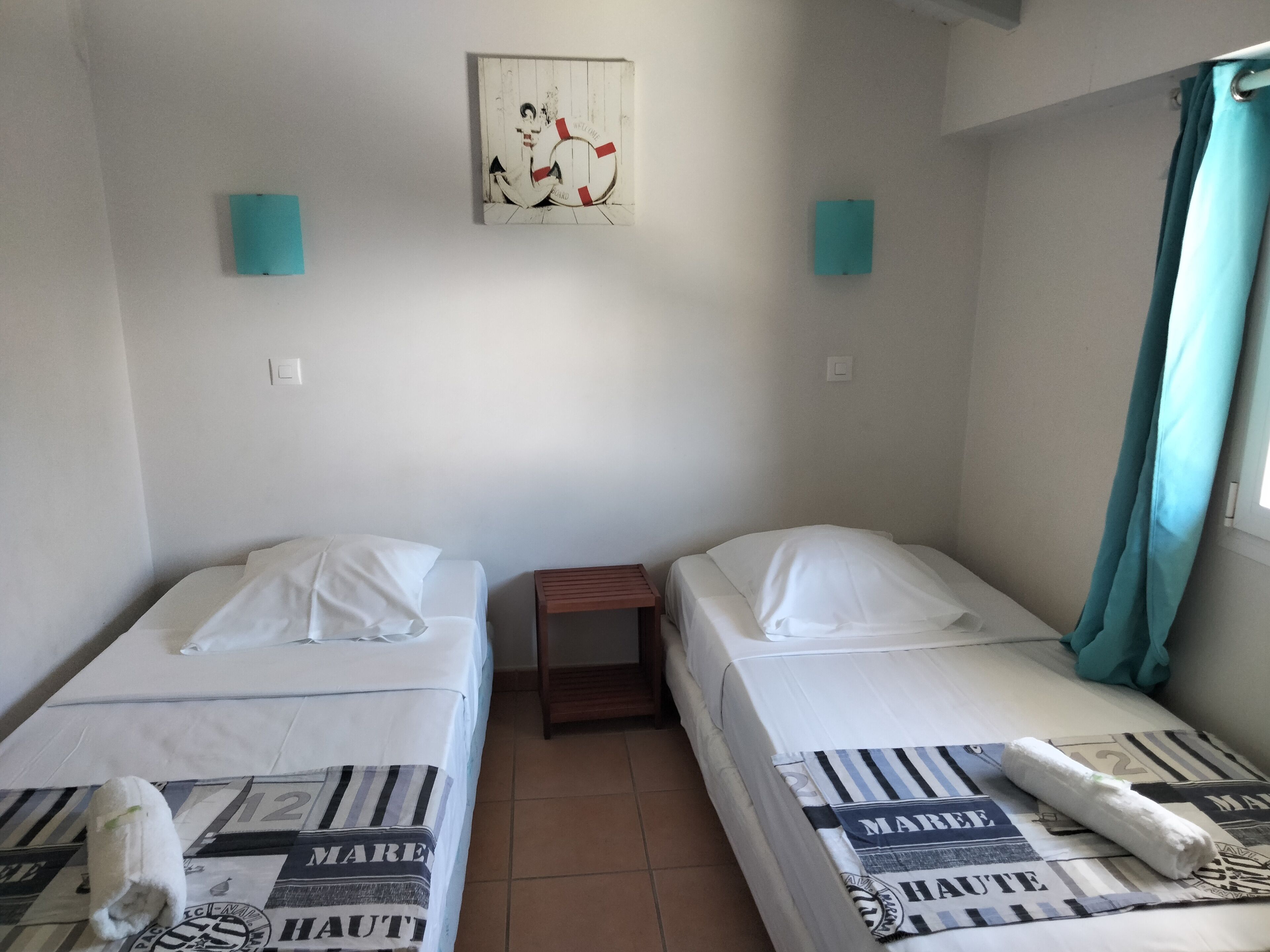 Une chambre avec deux lits simples, une petite table, une œuvre d’art accrochée au mur et des rideaux de couleur turquoise.