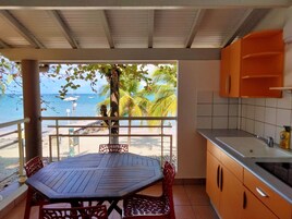 Un balcón con una mesa y sillas de madera, vista a la playa y al océano, y una cocina con gabinetes color naranja.