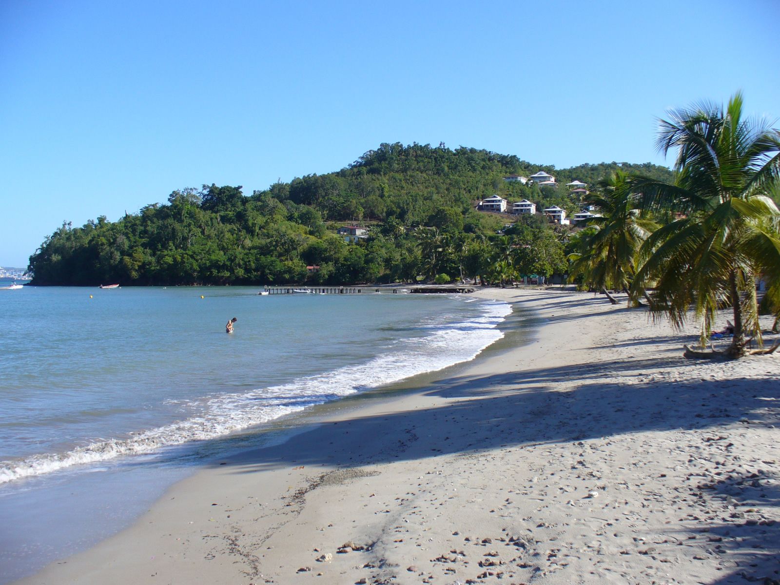 A beach with a clear view of the ocean, a sandy shore, and a hill with buildings in the background.