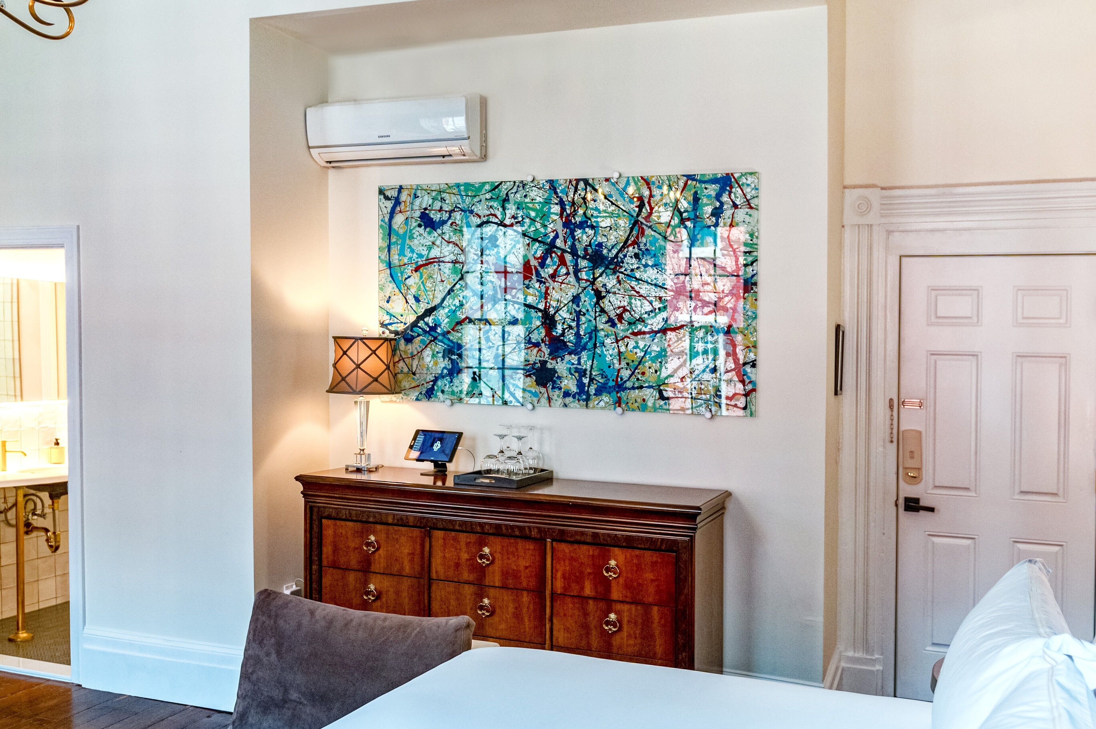 Una habitación de hotel con un cuadro abstracto grande en la pared, un aparador de madera, una cama y una puerta.