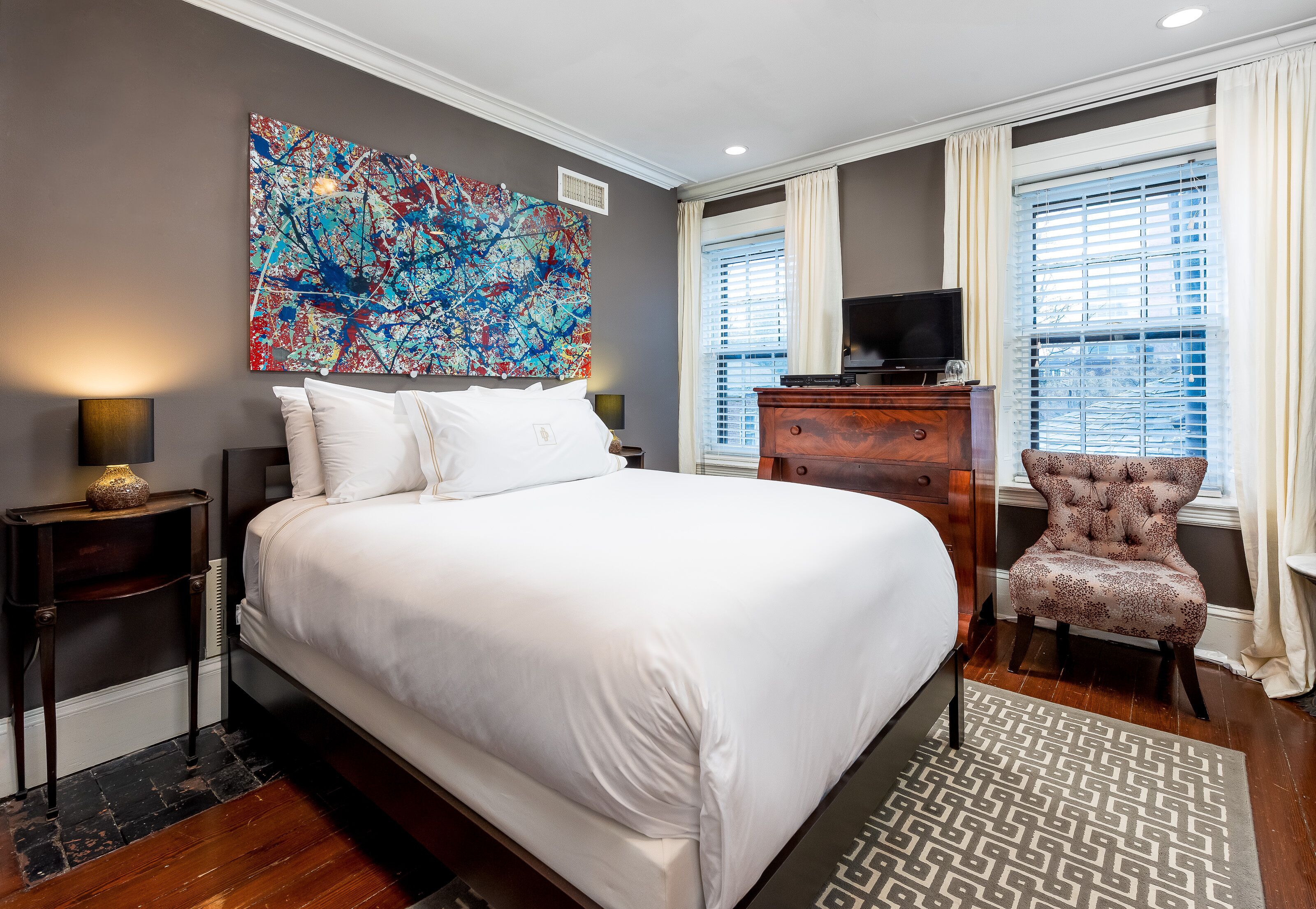 A modern hotel room with a large bed, a television, a decorative chair, and a large abstract painting on the wall.