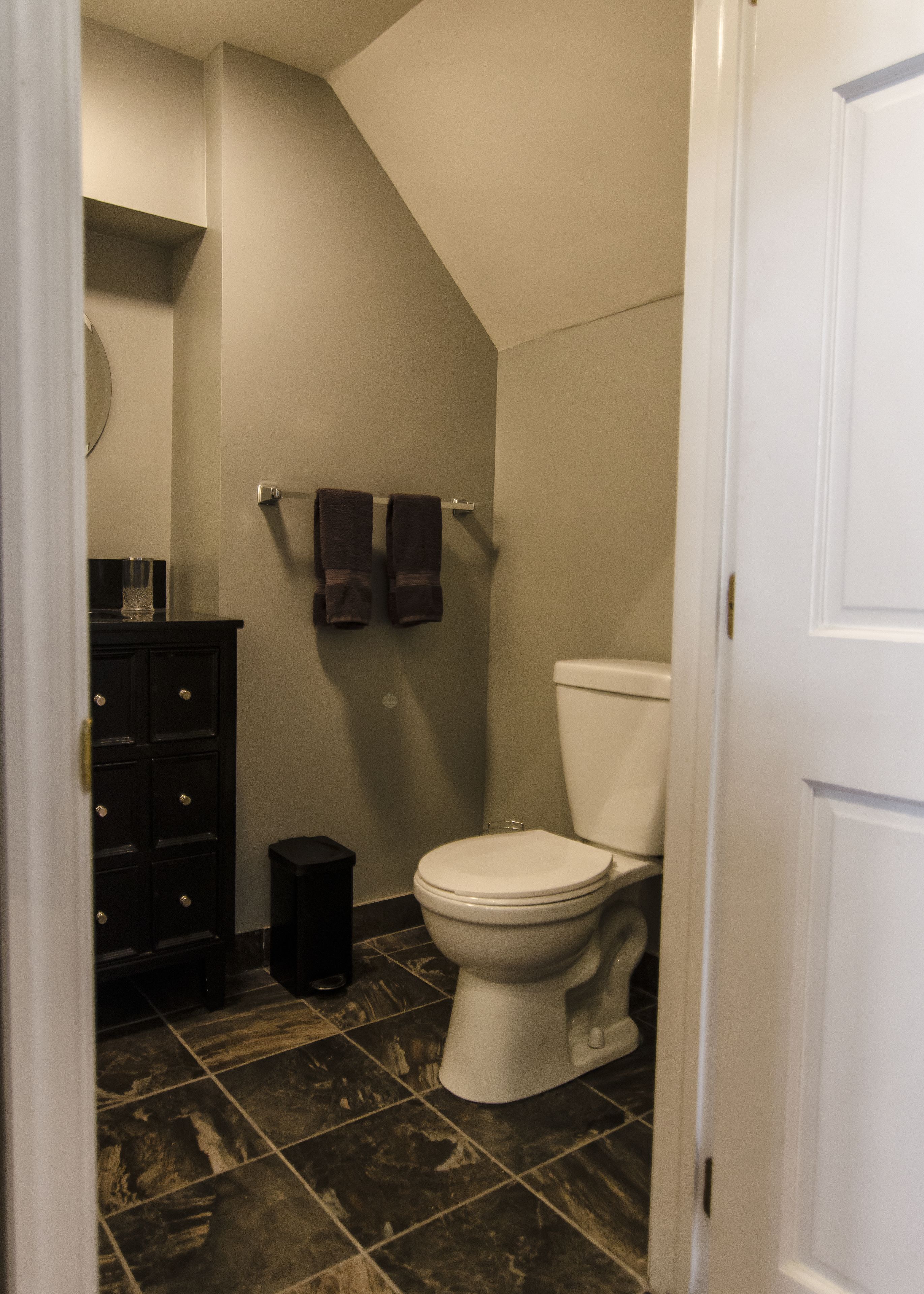 A bathroom with a toilet, dark tiled flooring, a dark cabinet, and two towels hanging on a towel rack.