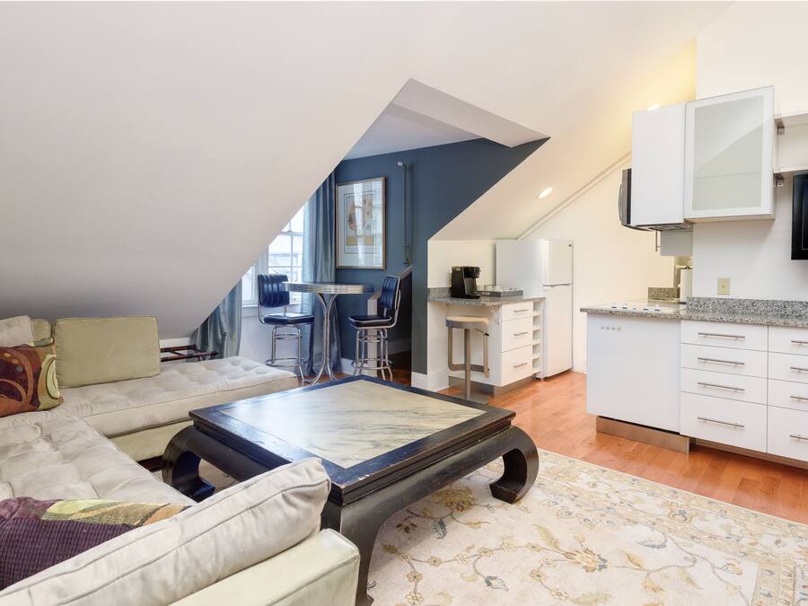 A modern living room with a sofa, coffee table, and a dining area with chairs and a kitchen with white cabinets and a TV.