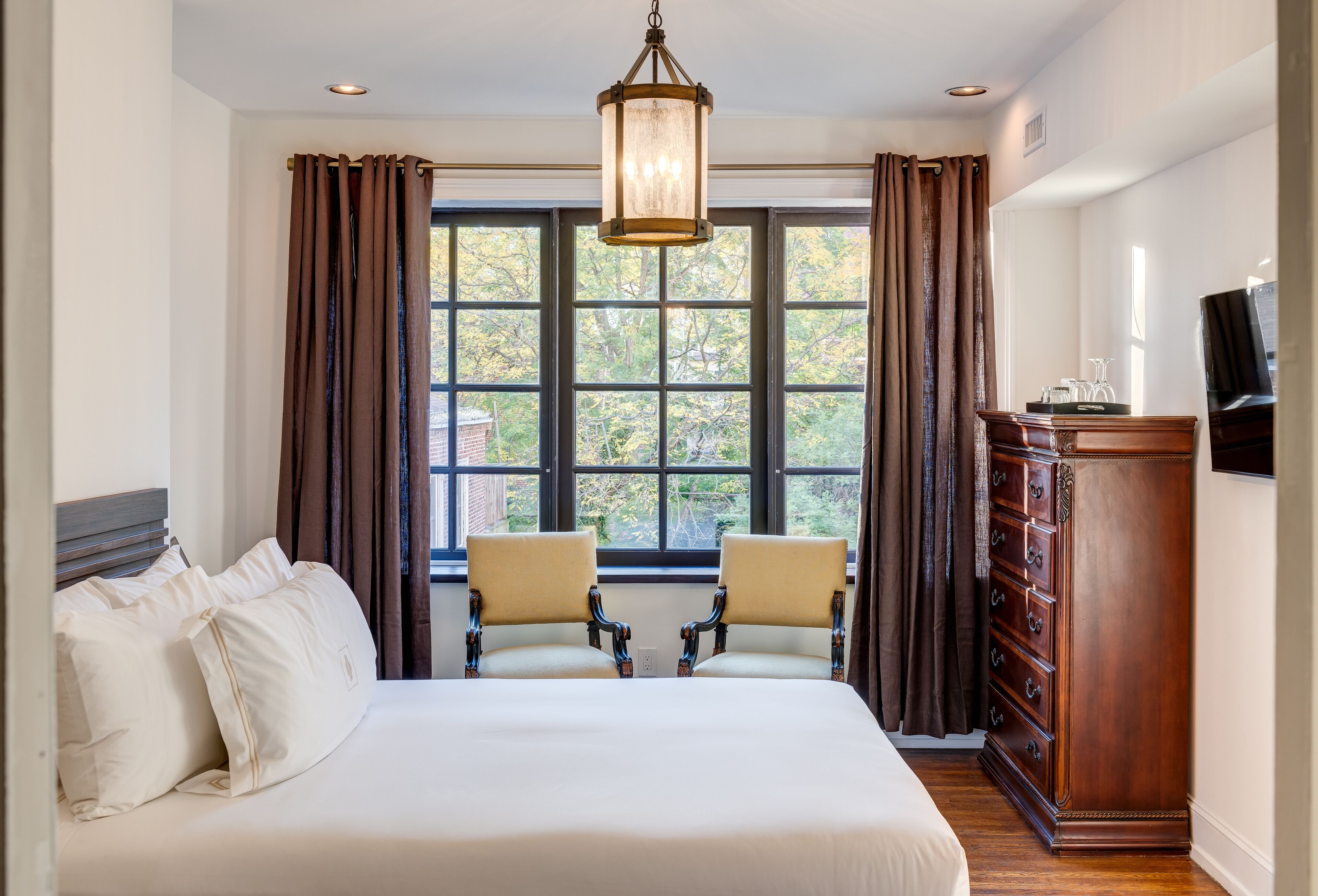 Een hotelkamer met een groot bed, twee fauteuils, een houten dressoir en een raam met gordijnen.