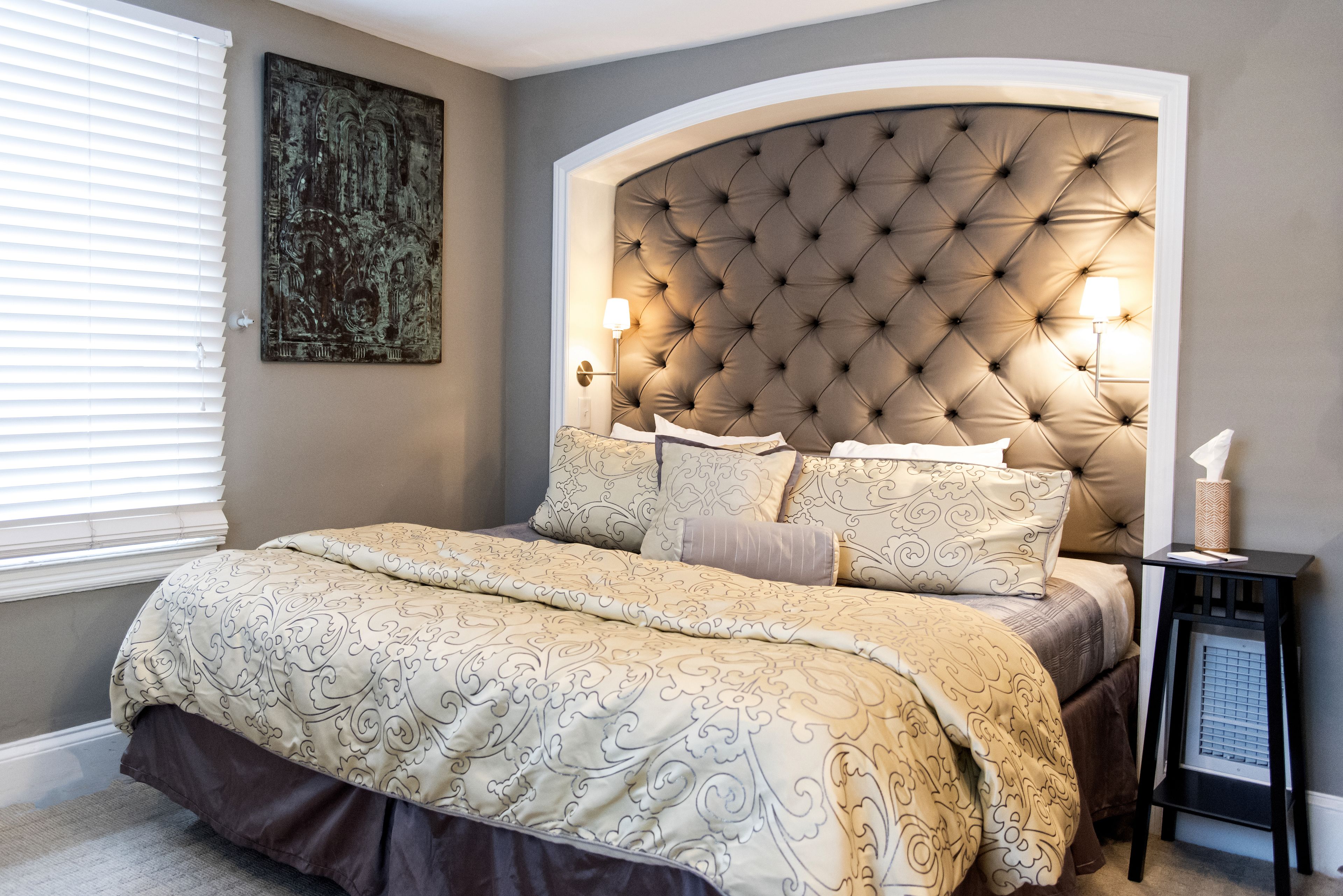 A bedroom with a tufted headboard, a large bed with patterned bedding, a bedside table with a lamp, and a wall-mounted artwork.