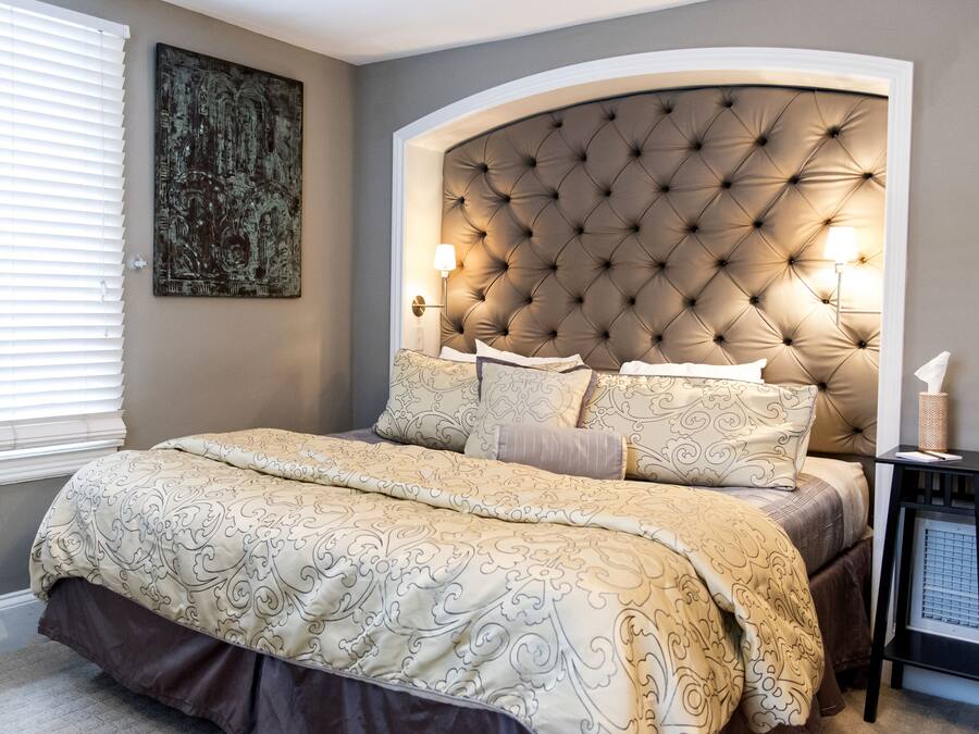 A bedroom with a tufted headboard, a large bed with patterned bedding, a bedside table with a lamp, and a wall-mounted artwork.