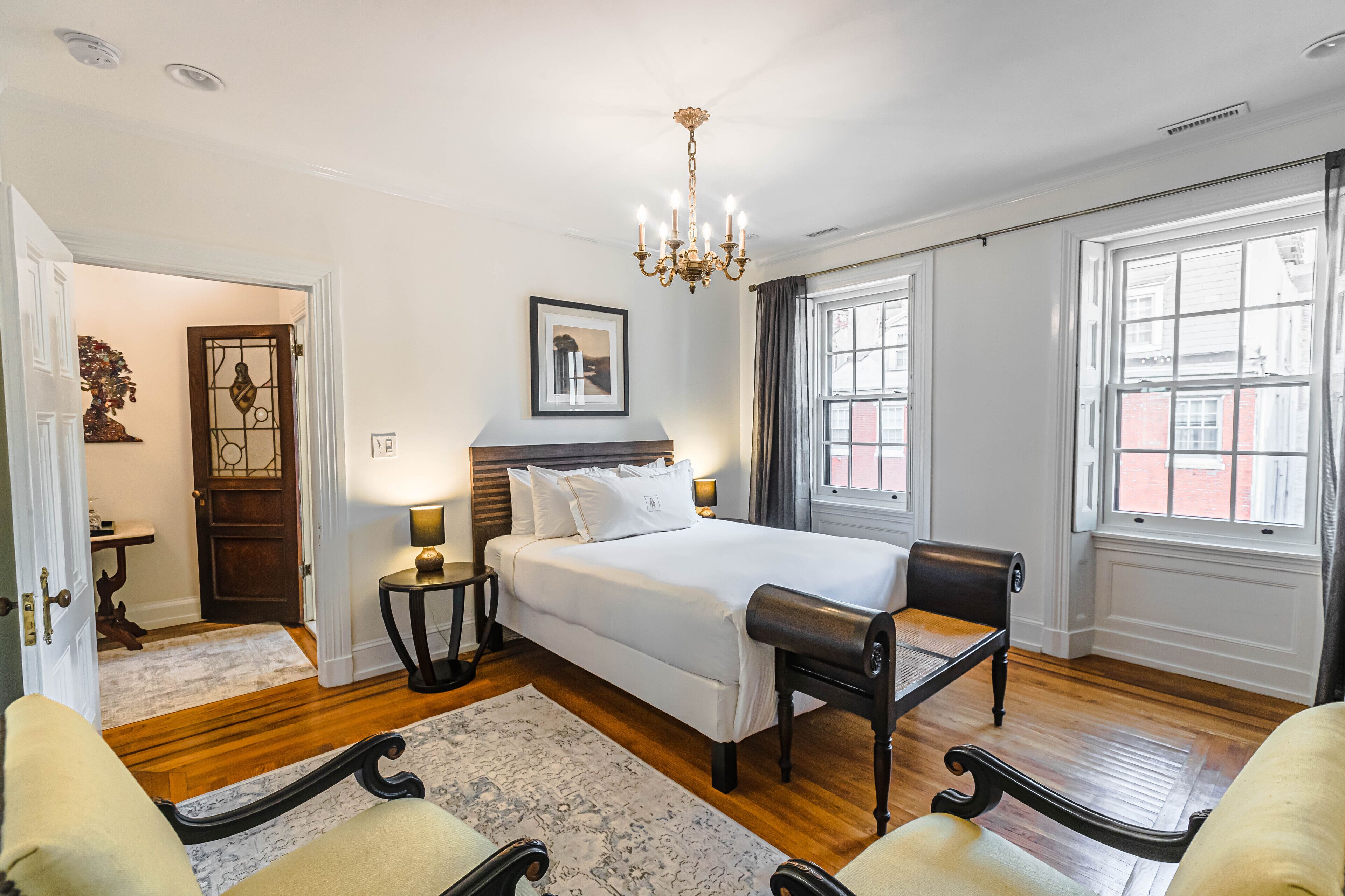 A spacious bedroom with a large bed, a sitting bench, a chandelier, and a view of the street through the windows.