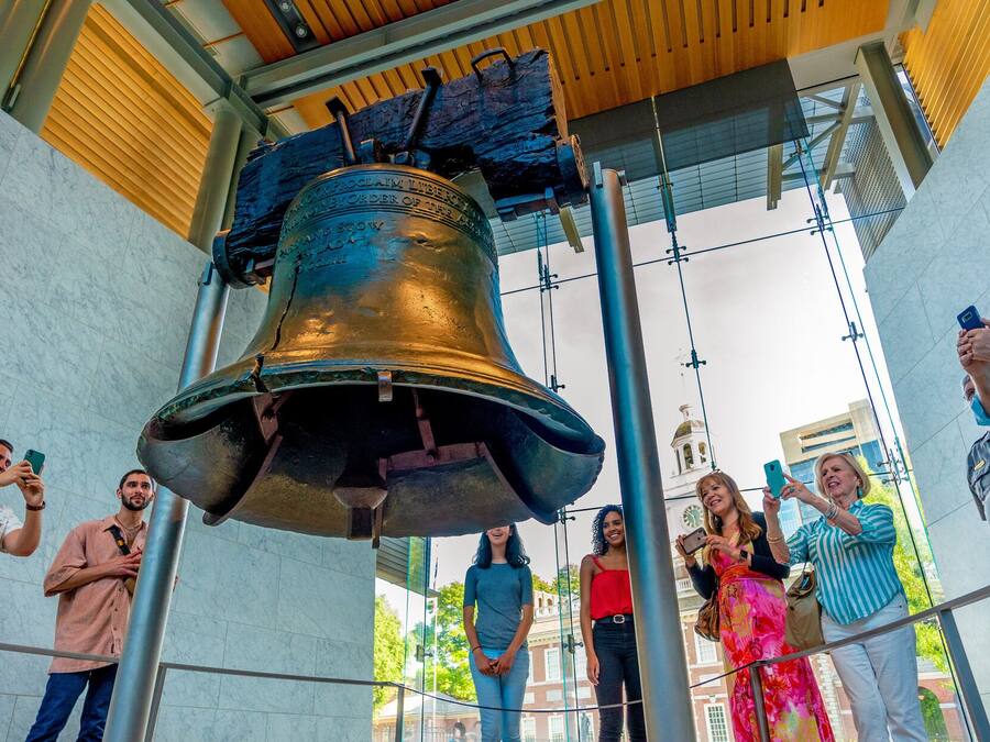 Eine große Glocke, aufgehängt in einem modernen Gebäude mit Glasfassade, umgeben von Menschen, die Fotos machen.