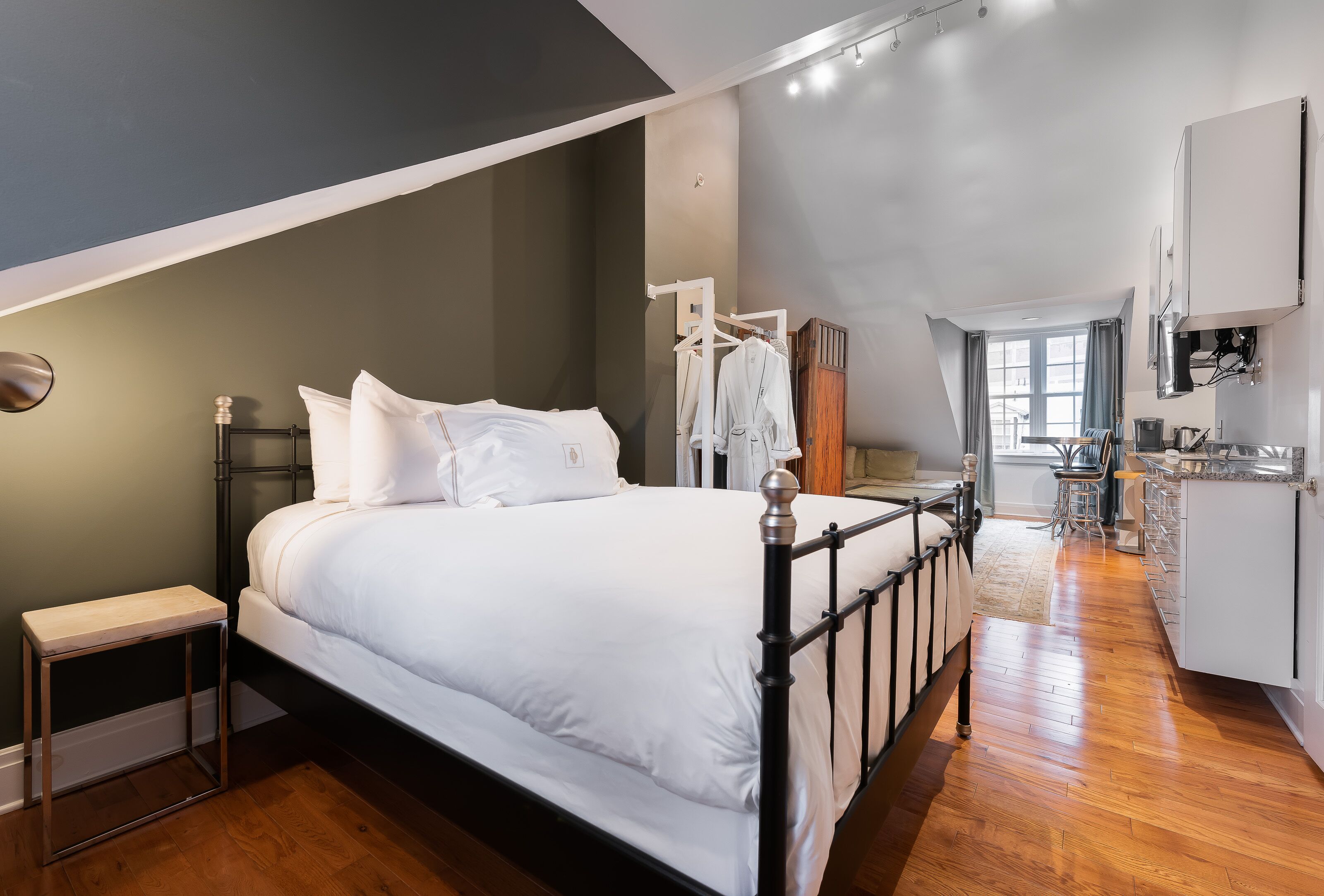 Een slaapkamer met een bed, nachtkastje en een klein krukje. Er is een kitchenette met een fornuis en kastjes.