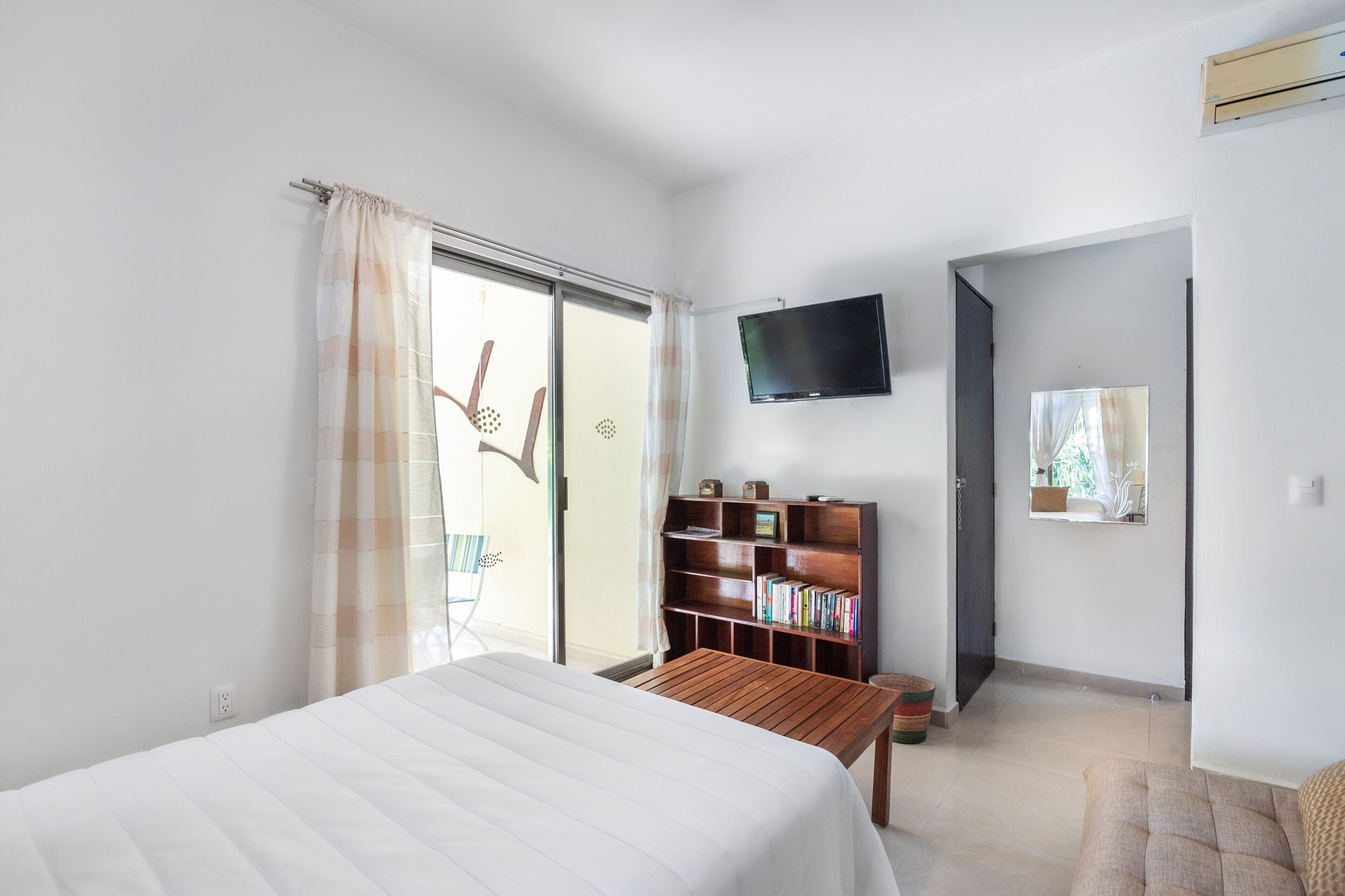 Quarto com uma cama, uma mesa de madeira, uma estante, uma televisão e uma porta de vidro deslizante.