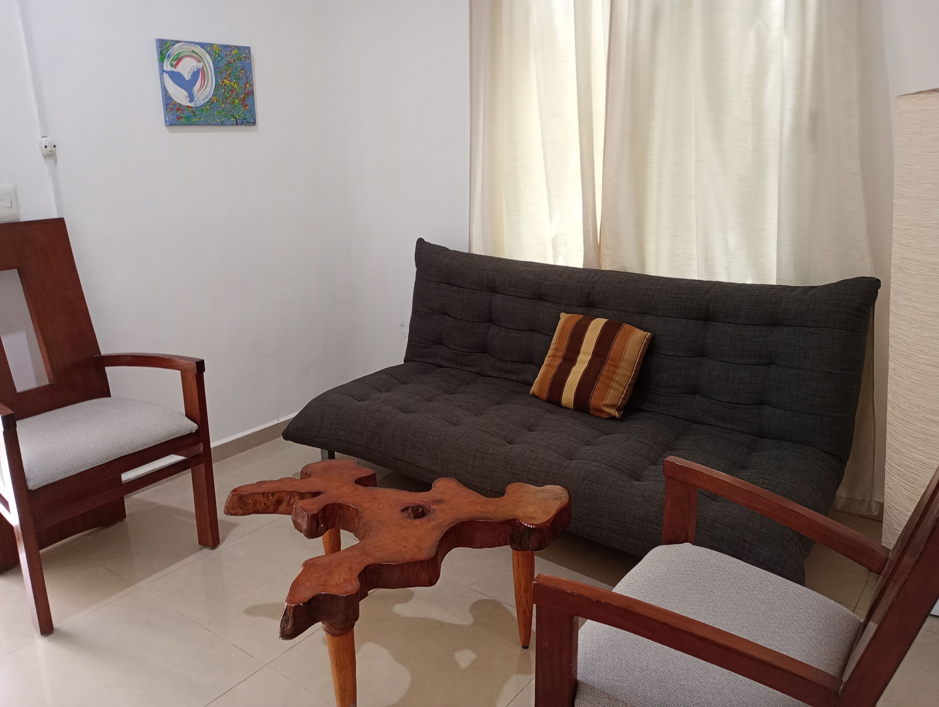 Un salon avec un canapé gris, deux fauteuils en bois, une table basse en bois et un tableau accroché au mur.