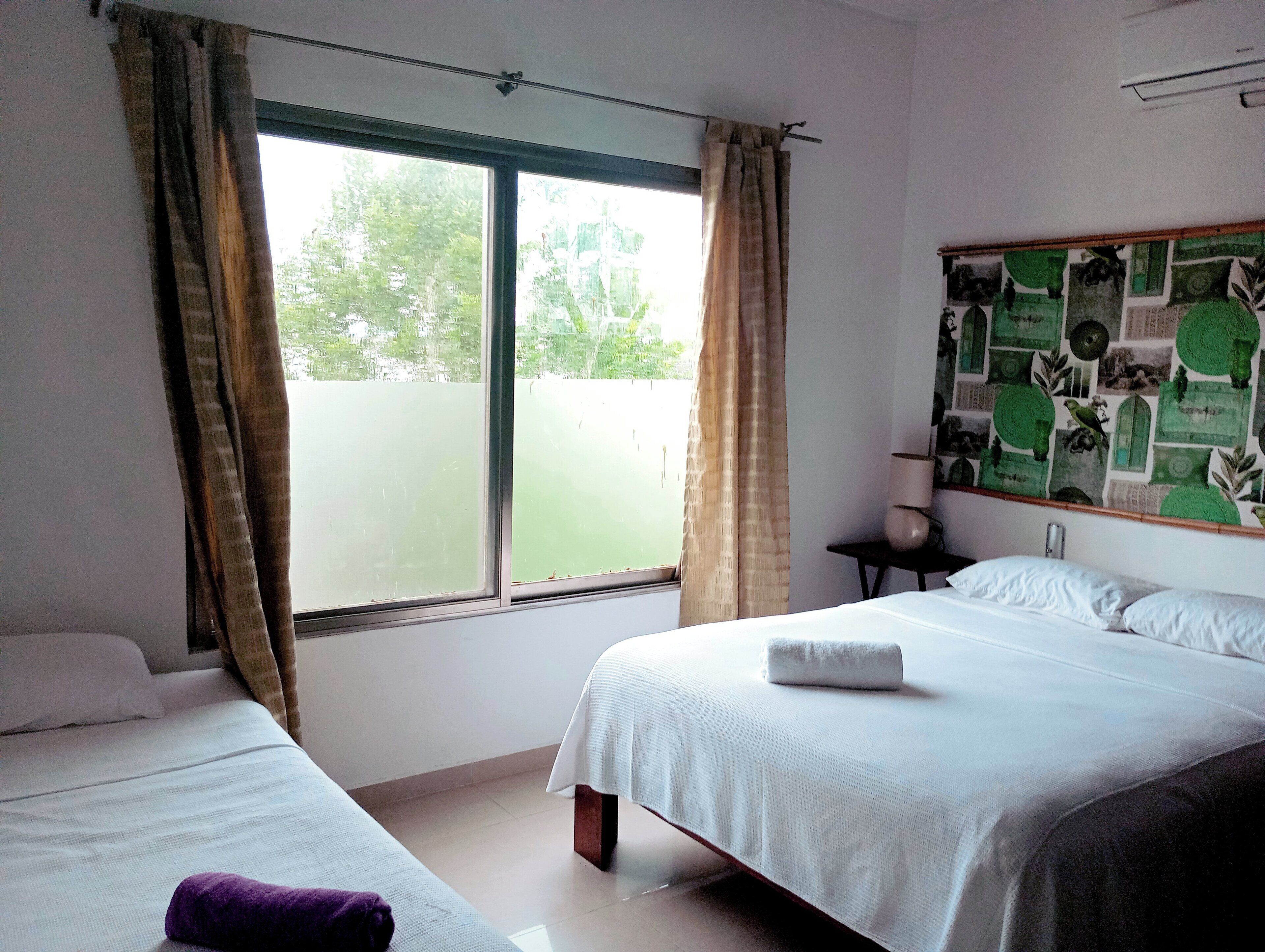 A hotel room with two beds, a window with curtains, a wall with a green and white patterned mural, and a small table with a lamp.