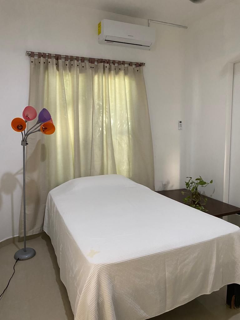 A bedroom with a bed, a standing lamp, a potted plant, and an air conditioning unit.