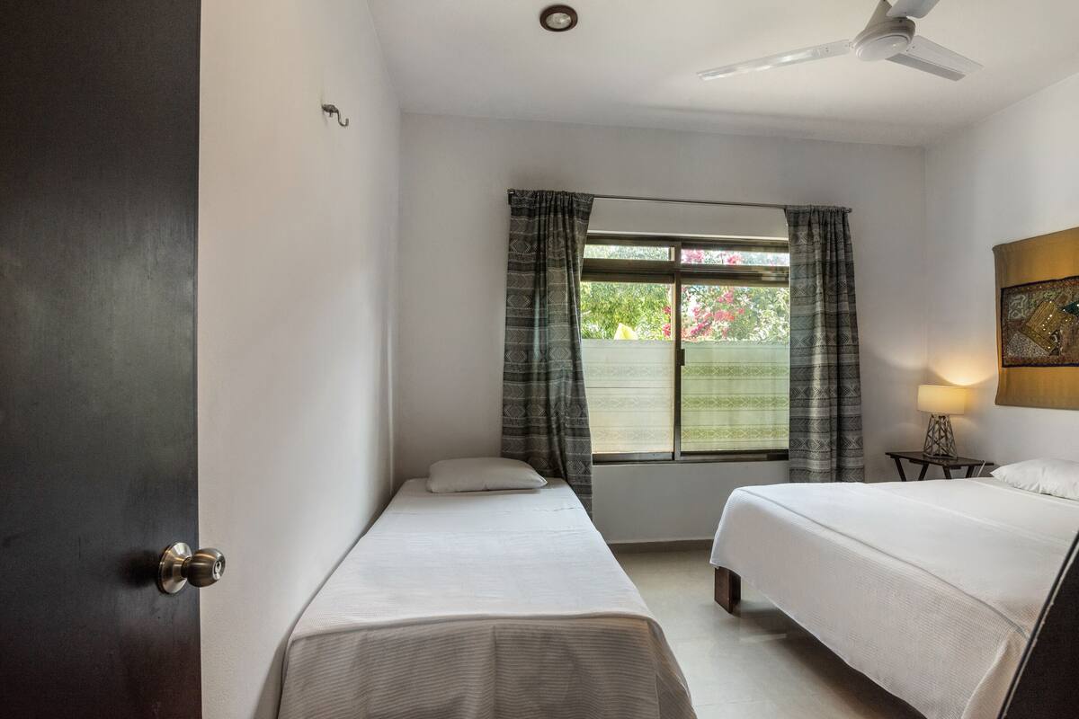Ein Zimmer mit zwei Einzelbetten, einem Deckenventilator, einem Fenster mit Vorhängen und einem Nachttisch mit Lampe.