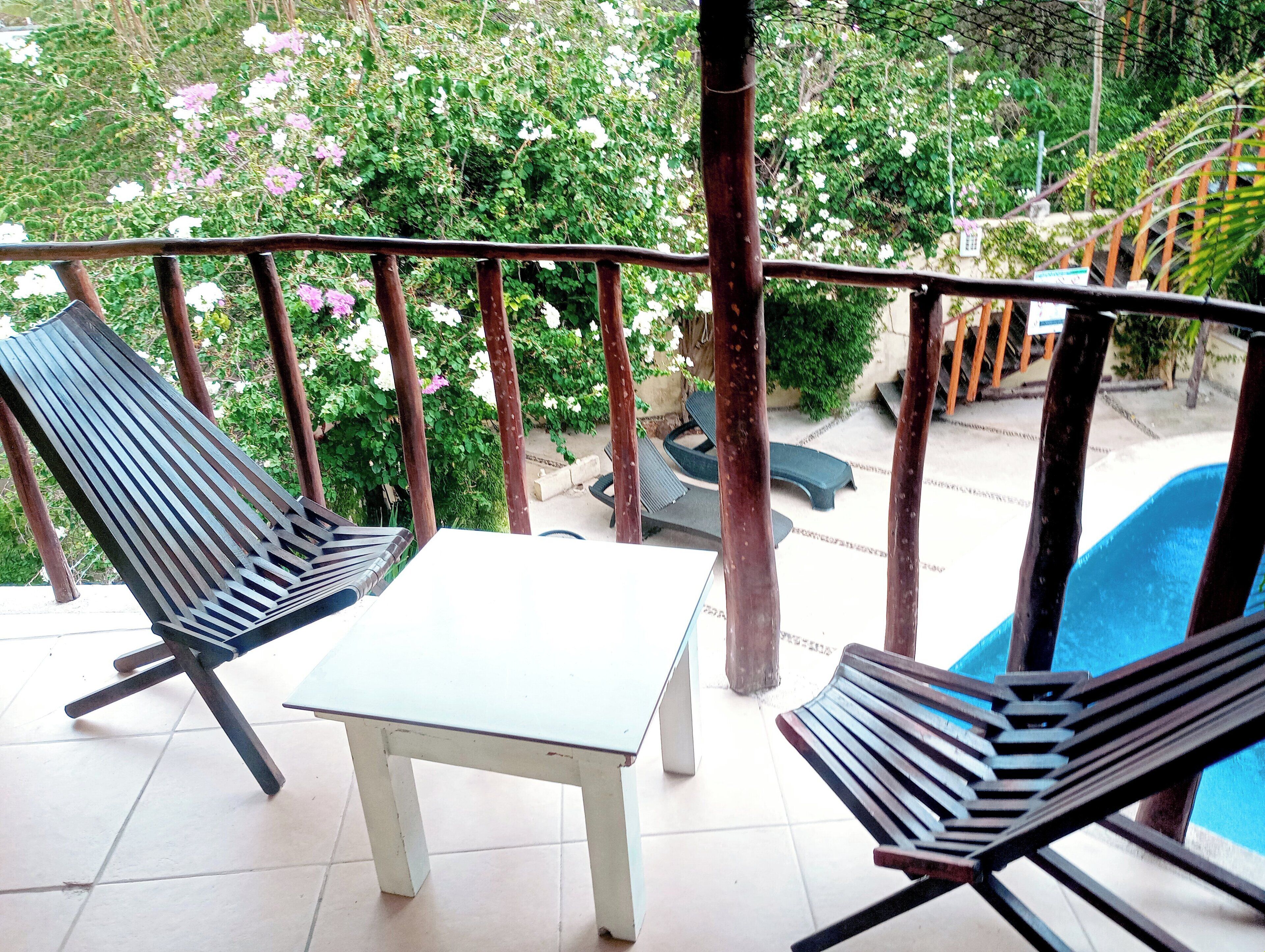 Ein Balkon mit zwei Holzliegestühlen und einem kleinen Tisch, mit Blick auf einen Swimmingpool und üppiges Grün.