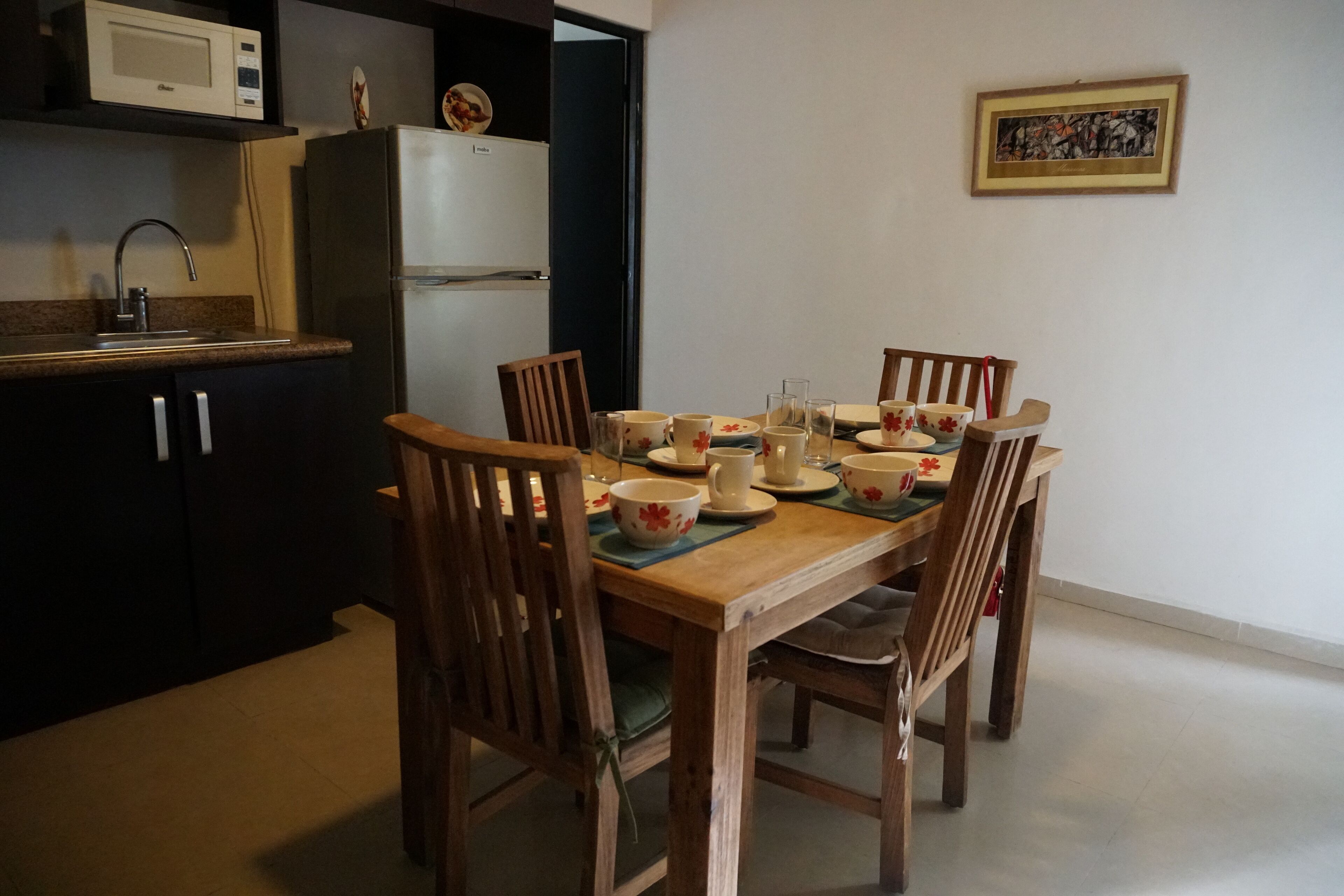 Un coin repas avec une table en bois dressée pour quatre personnes, entourée de chaises. On peut apercevoir un four à micro-ondes et un réfrigérateur à l’arrière-plan.
