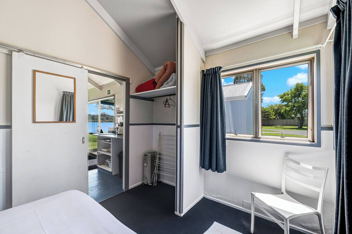 A compact hotel room with a bed, a white chair, a small desk, a radiator, and a window with curtains.