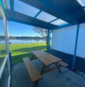 A covered outdoor area with a picnic table, a view of a lake, and a building in the background.