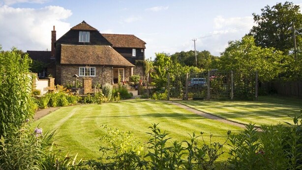 A two-story house with a stone facade, a well-maintained lawn, and a garden with various plants and flowers. - Magpies (Horsham)